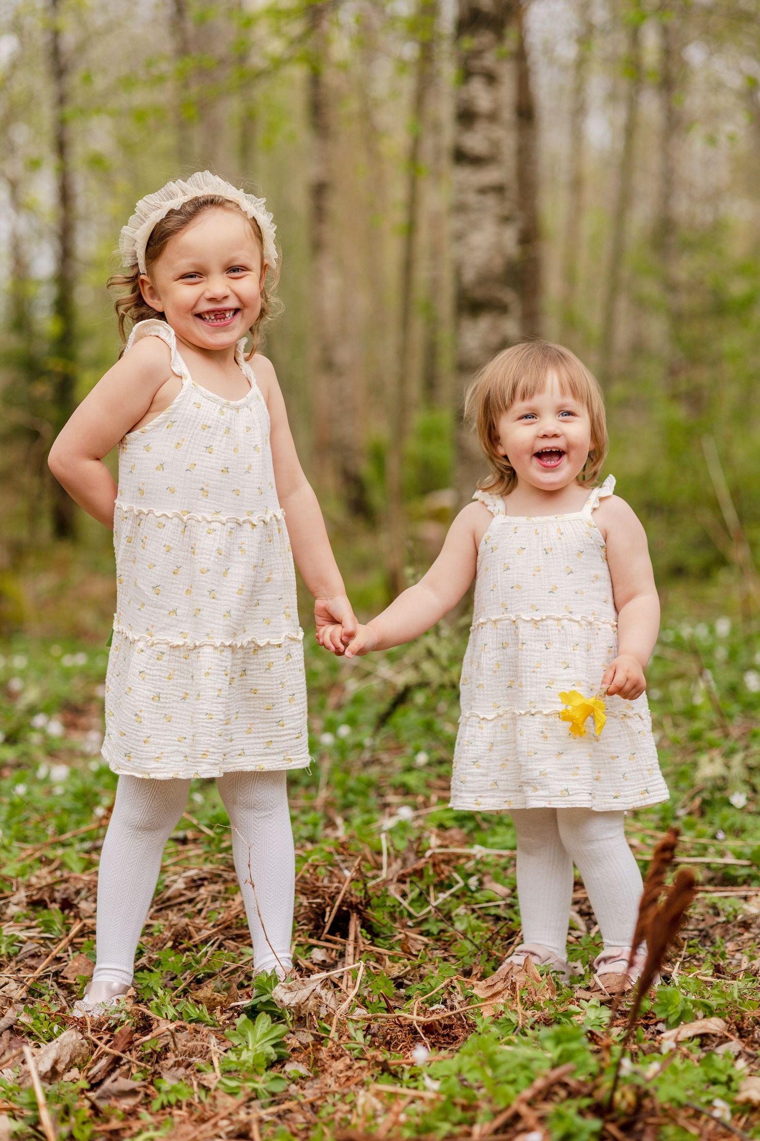 Two giggling little girls in matching cream floral dresses hold hands among green forest undergrowth and white spring flowers.