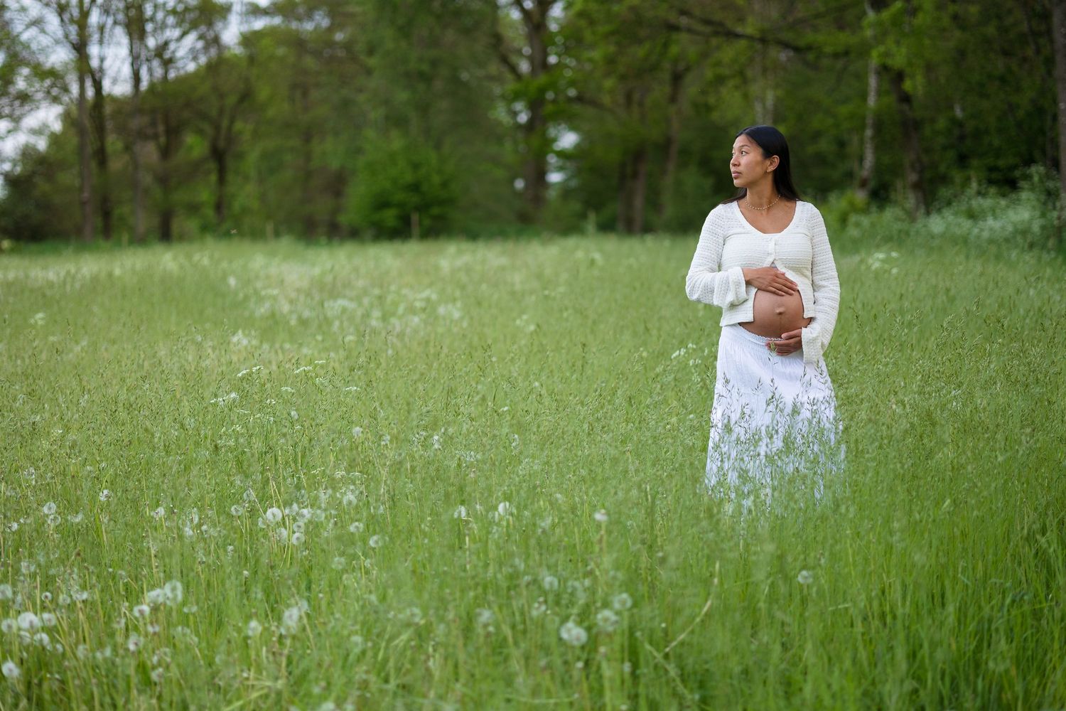 Pregnant woman in a white cardigan and skirt cradles her belly while standing in a green meadow with wildflowers.