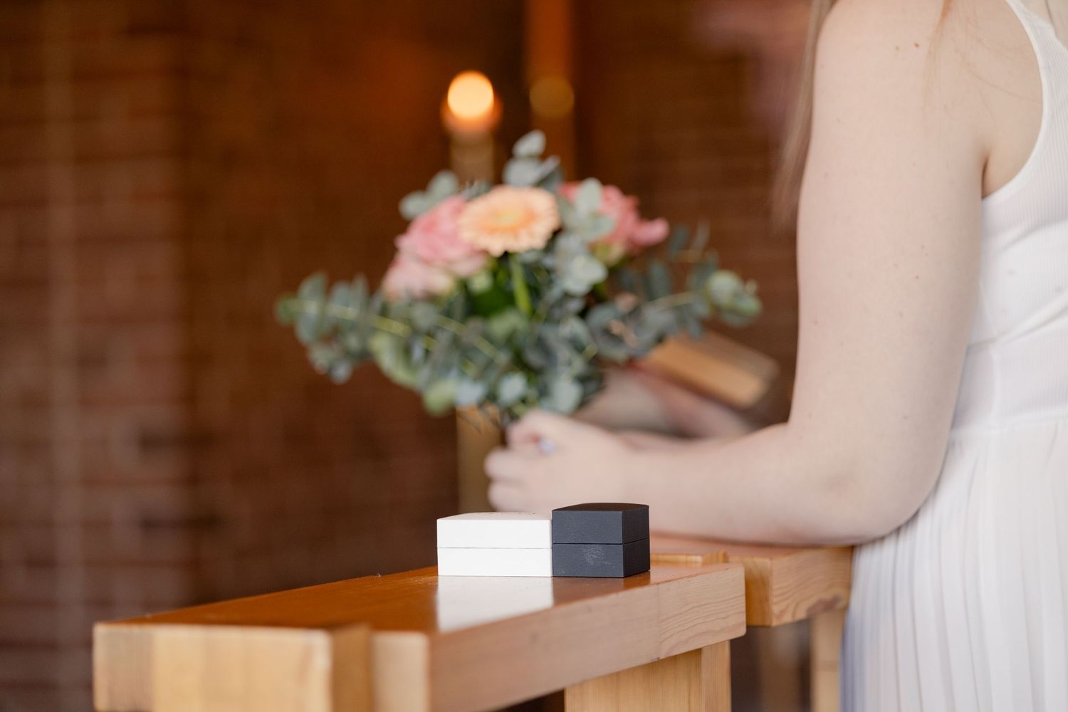 A woman in a white dress stands at a wooden table with two ring boxes and a flower arrangement in a warm candlelit room.