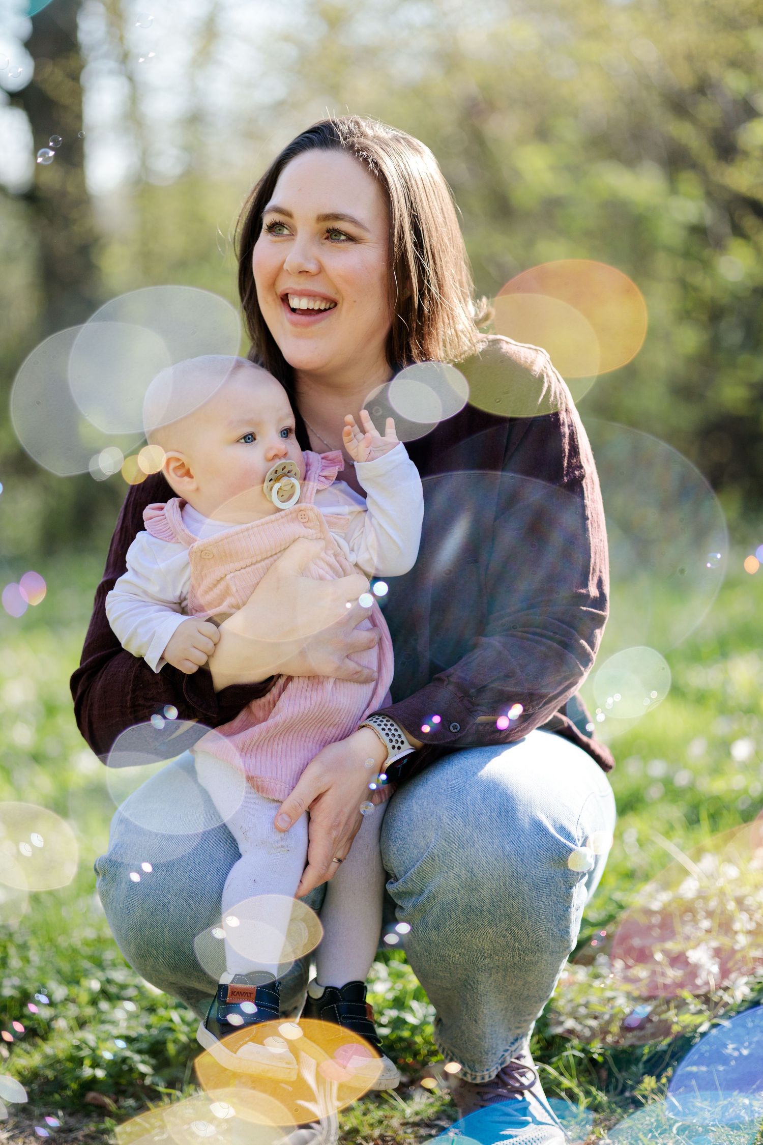 Laughing woman crouching outdoors with a baby in a pink outfit and pacifier while soap bubbles float around them.