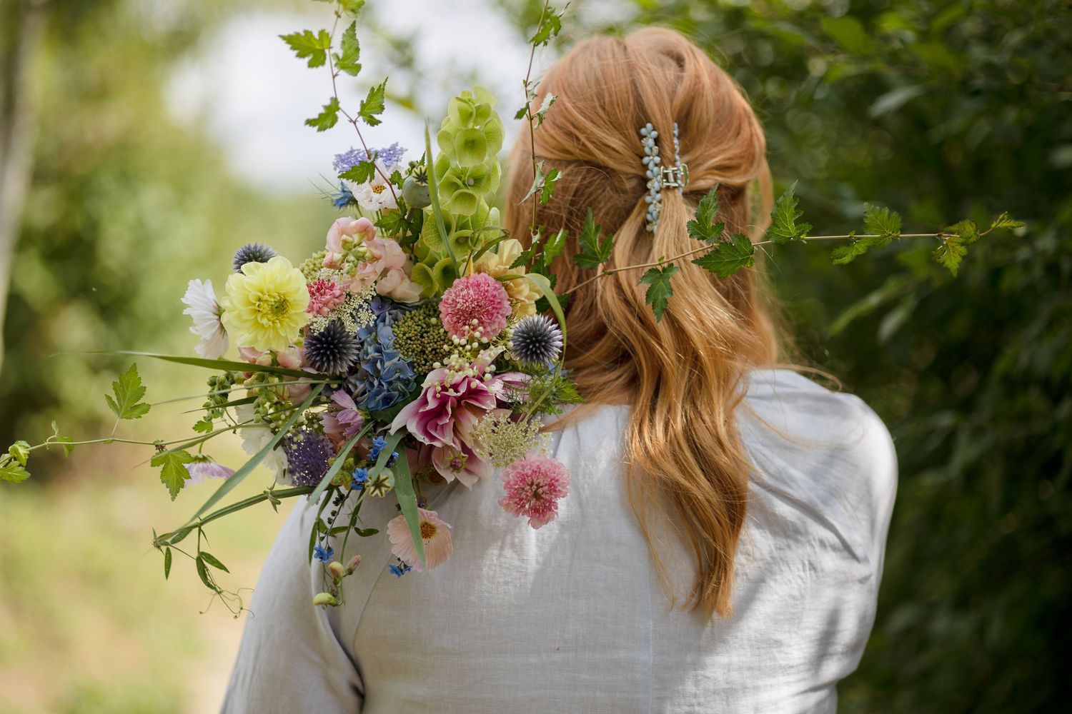 Woman with long auburn hair and a clip holding a colorful wildflower bouquet while facing a green garden.