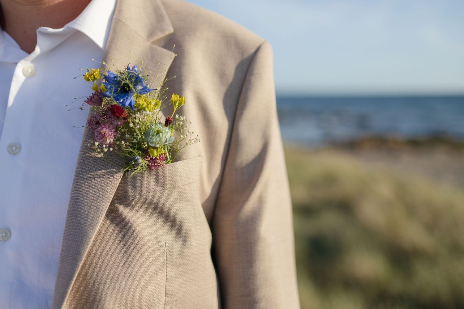 Close-up of a man's beige suit jacket with a colorful wildflower boutonniere beside a blurred seaside meadow.