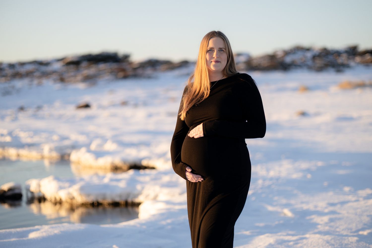 A pregnant woman in a black dress holds her baby bump on a snowy coast in warm golden evening light.
