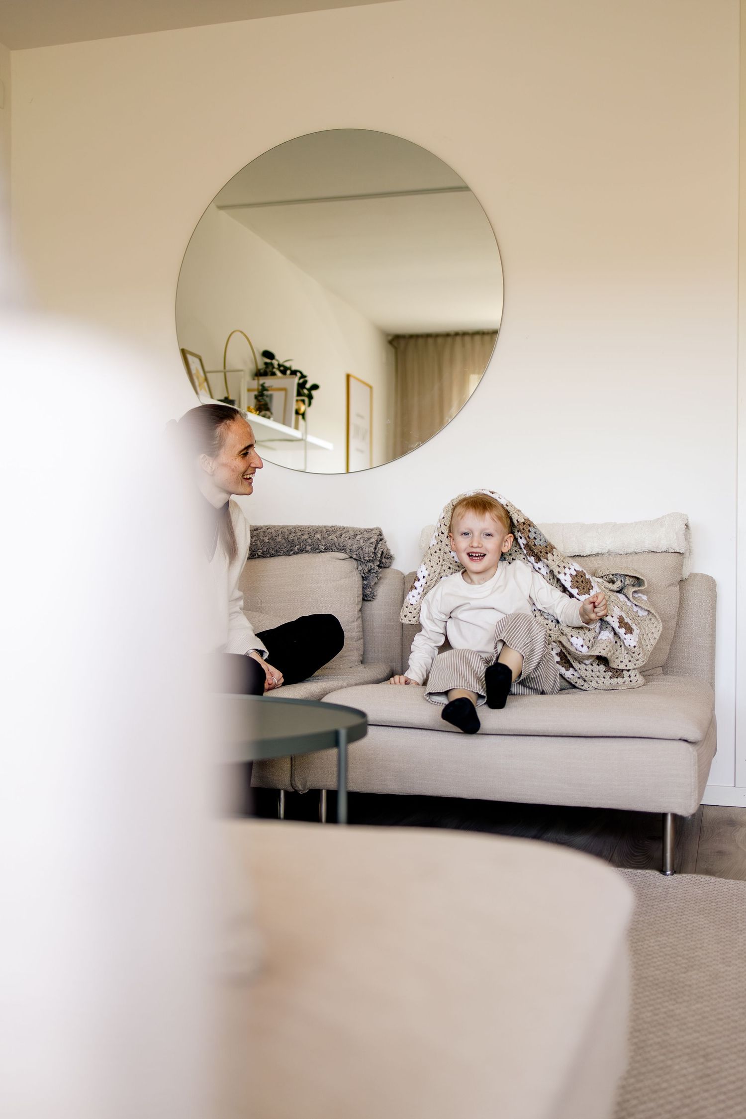 Smiling toddler wrapped in a patterned blanket sits on a beige sofa beside a woman, with a round mirror on the wall behind.