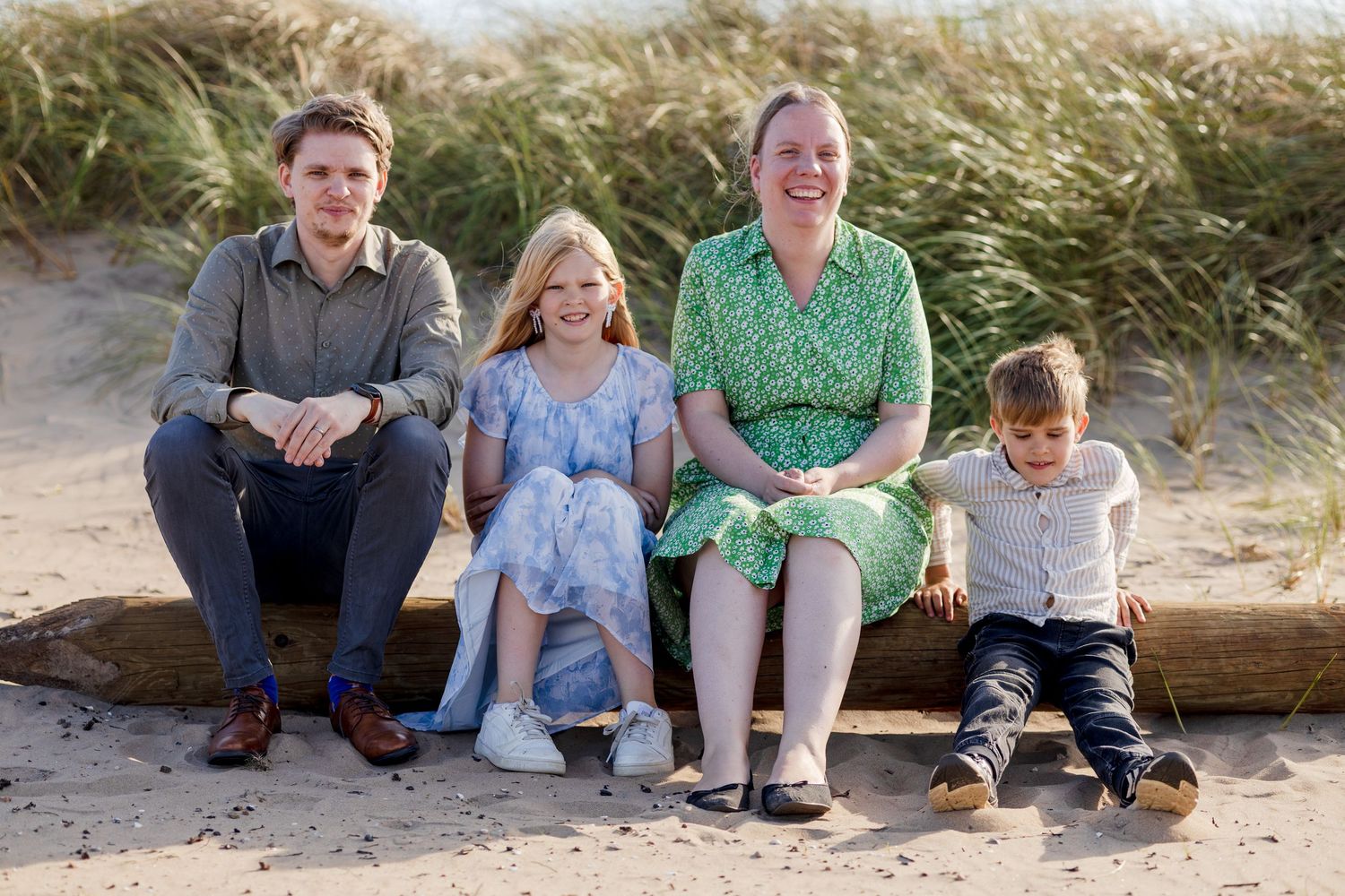 A family of four sits on a log at a sandy beach with tall dune grass, smiling together during a sunny outdoor portrait session.