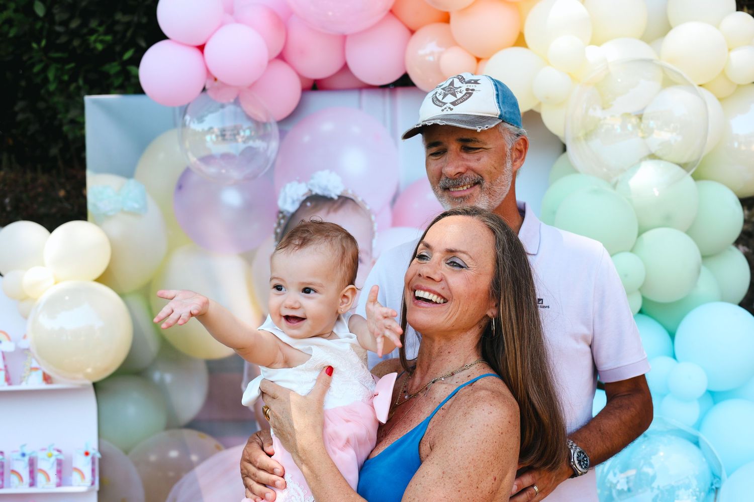Family posing with cheerful baby at colorful pastel balloon arch birthday party outdoors.