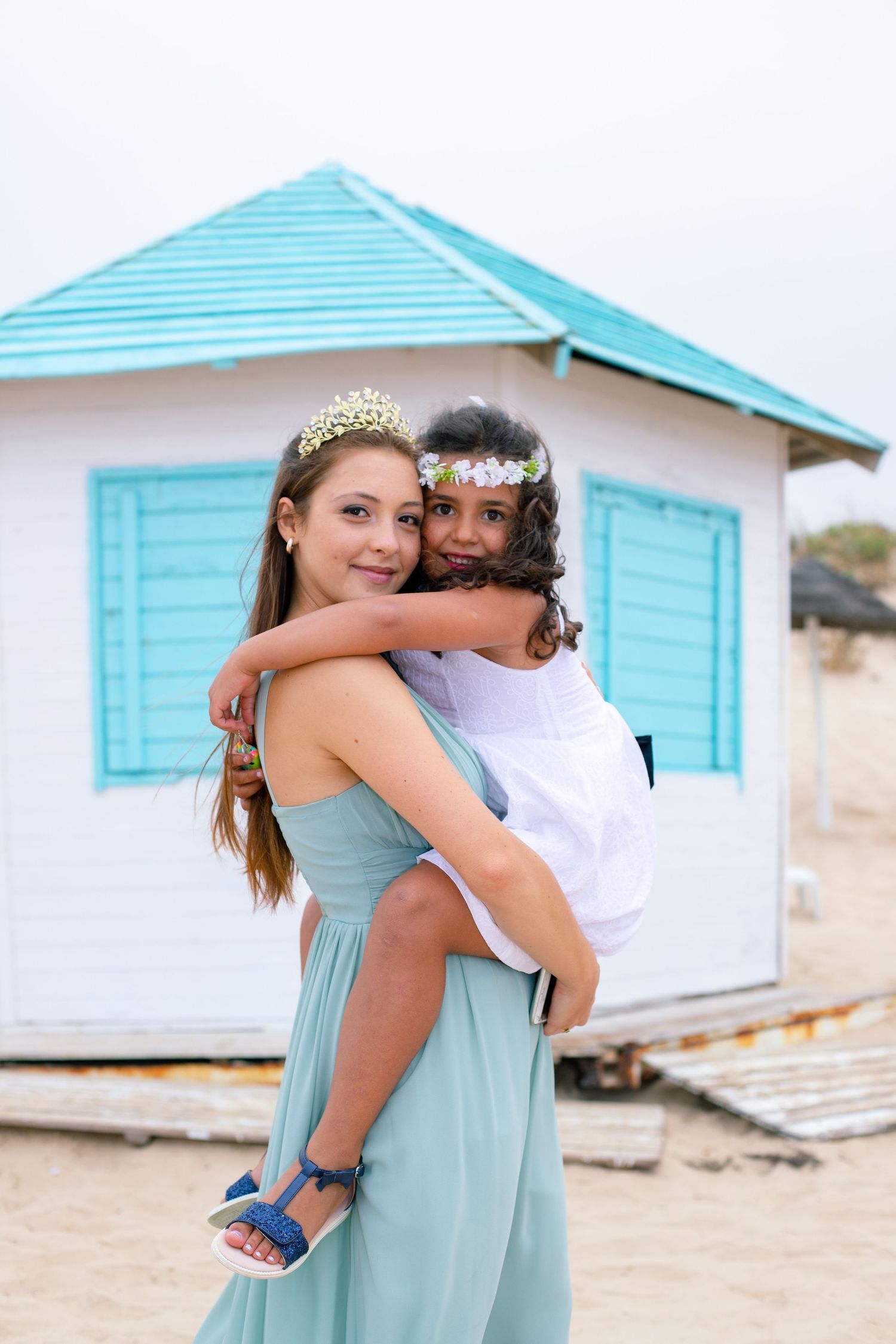 Young woman in teal dress holding a little girl wearing a flower crown near a beach hut.