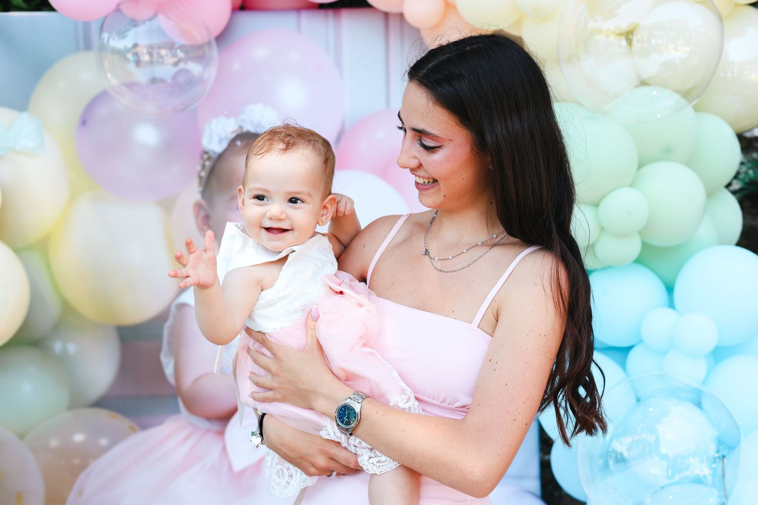 Young mother in pink dress smiling while holding her baby girl at a pastel balloon birthday party.
