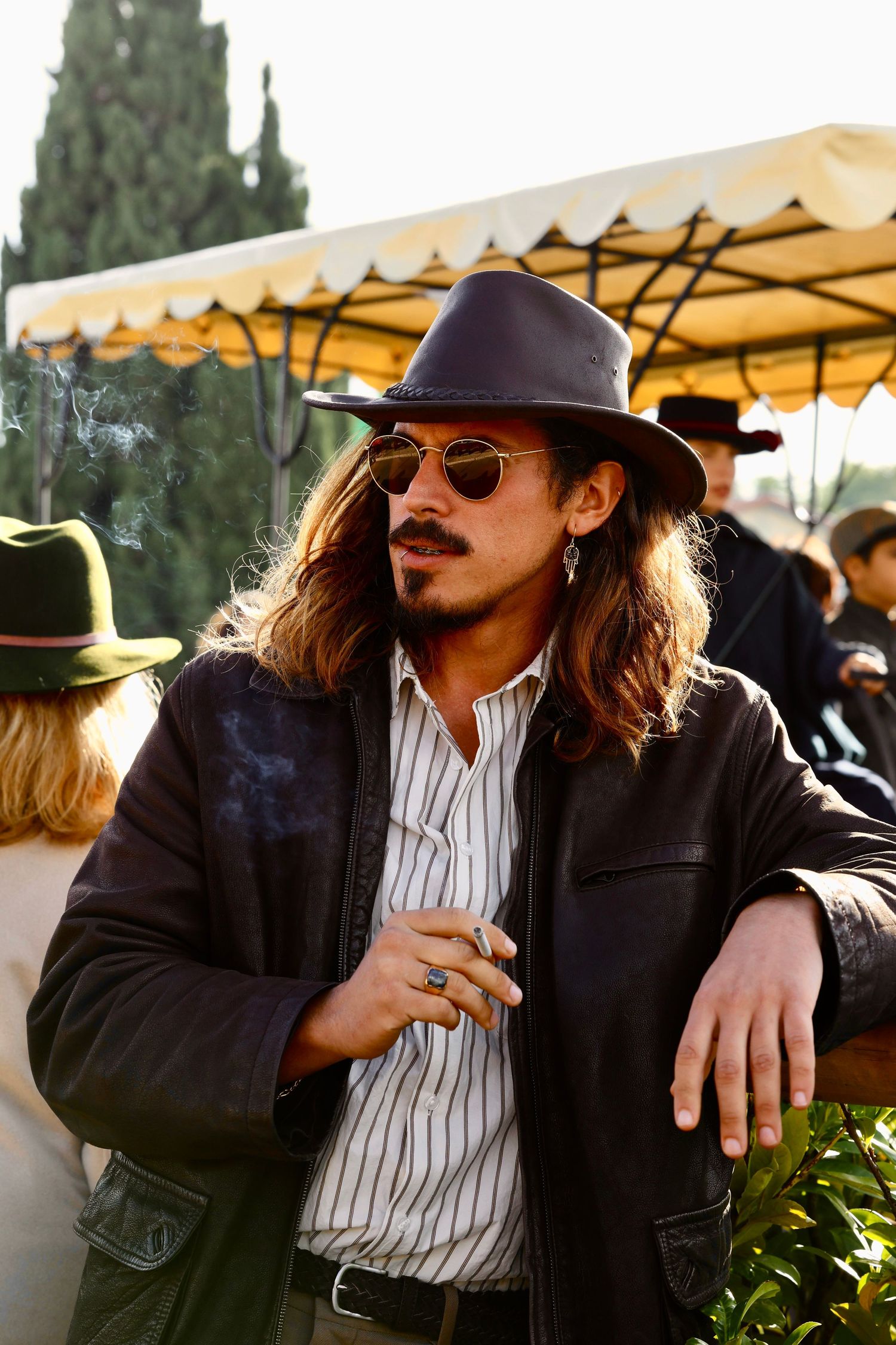 Stylish man with long hair wearing fedora and sunglasses smoking a cigarette outdoors.