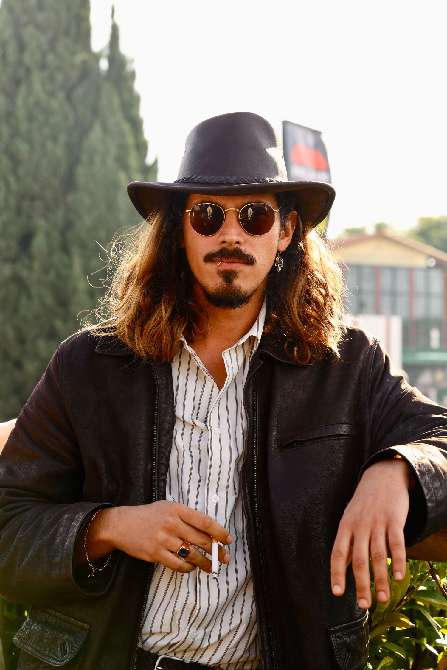 Fashionable man with long hair, fedora, round sunglasses and leather jacket posing outdoors.