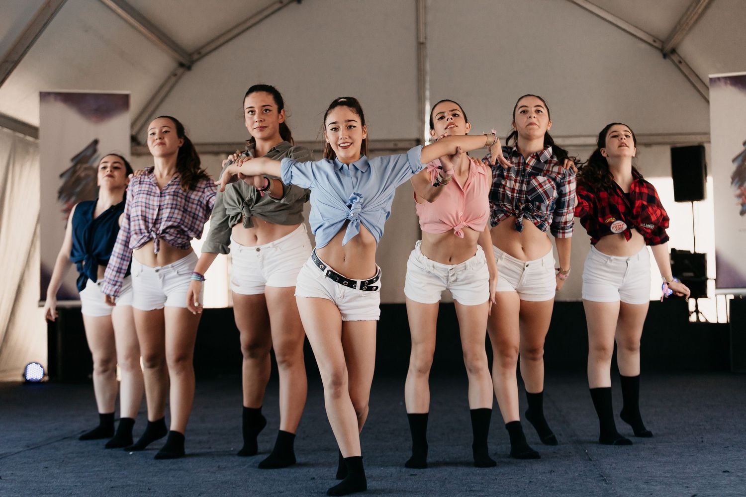 Seven dancers in plaid shirts and white shorts perform a synchronized routine on an outdoor stage.