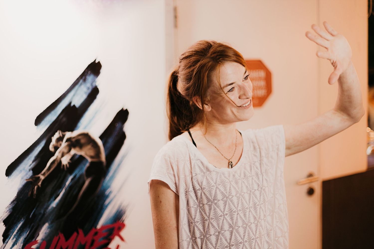A young woman with a ponytail smiles and waves her hand near a movie poster on a white wall.