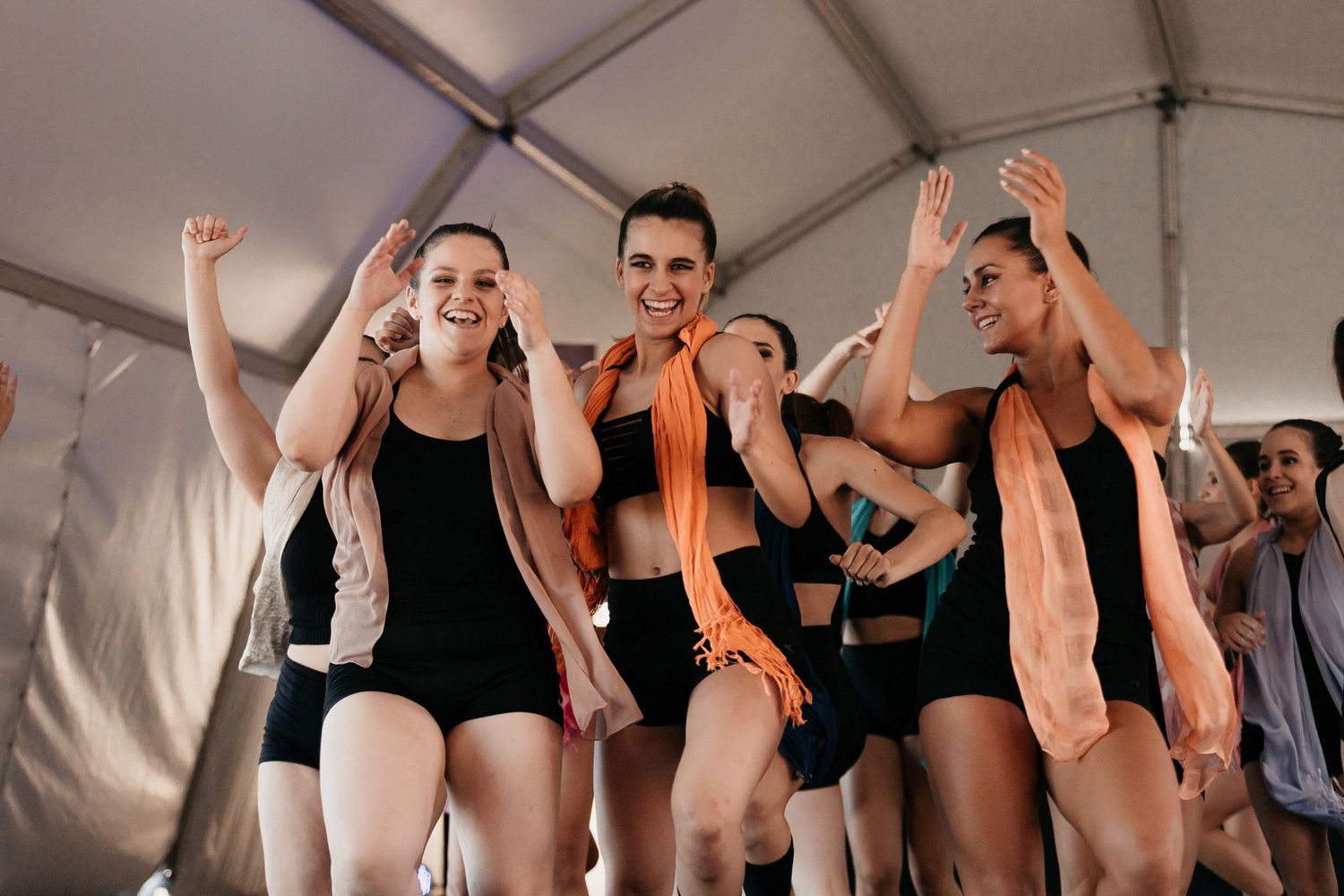 Dancers in black leotards celebrate joyfully under a white tent, wearing colorful orange scarves.
