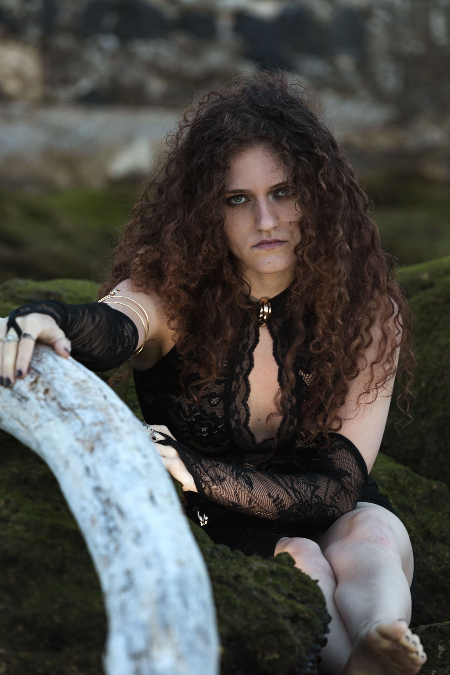 Woman with curly hair in black lace outfit posing on mossy rocks outdoors.