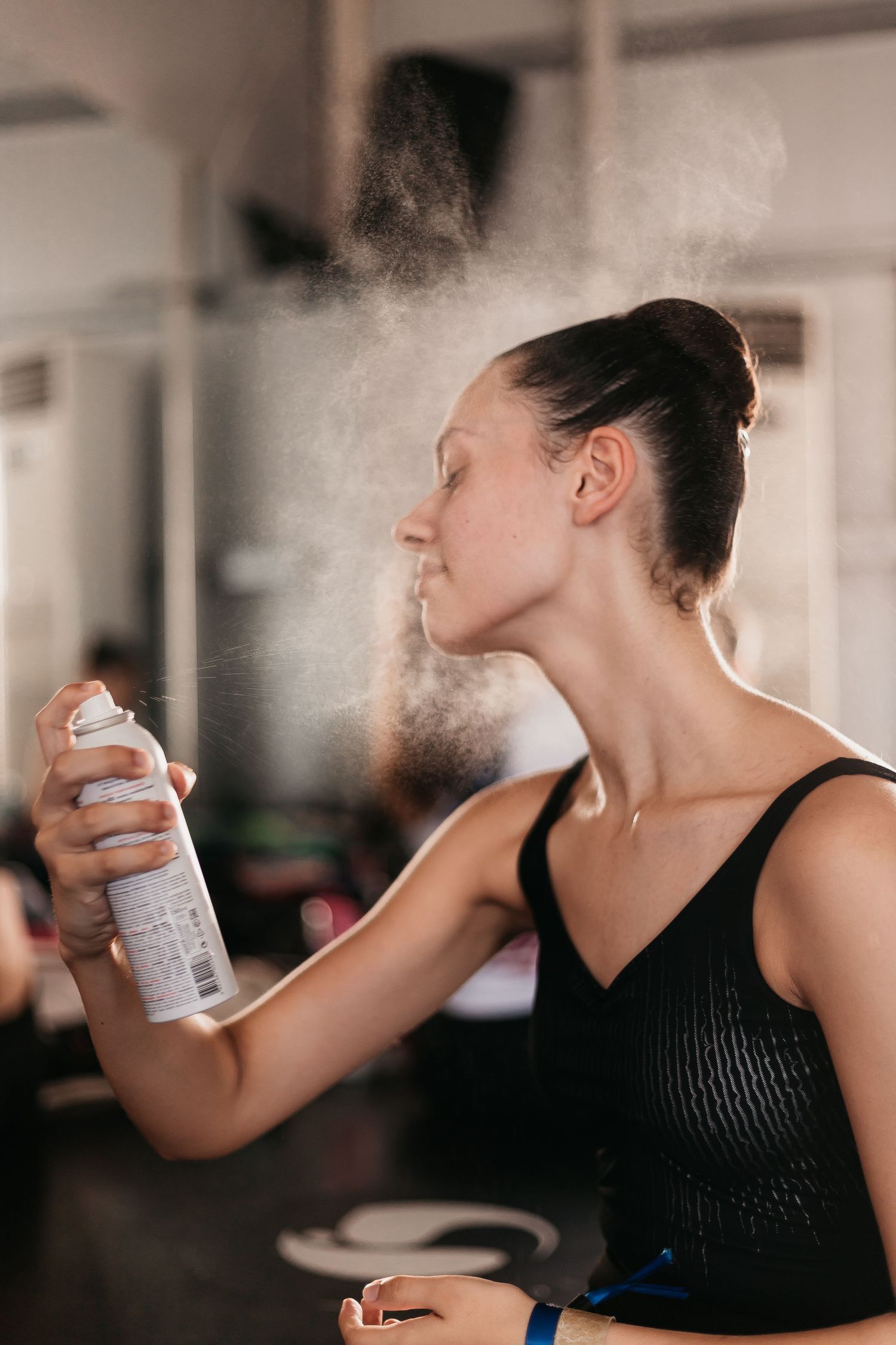 A ballet dancer in a black leotard sprays setting spray on her face in a dressing room.