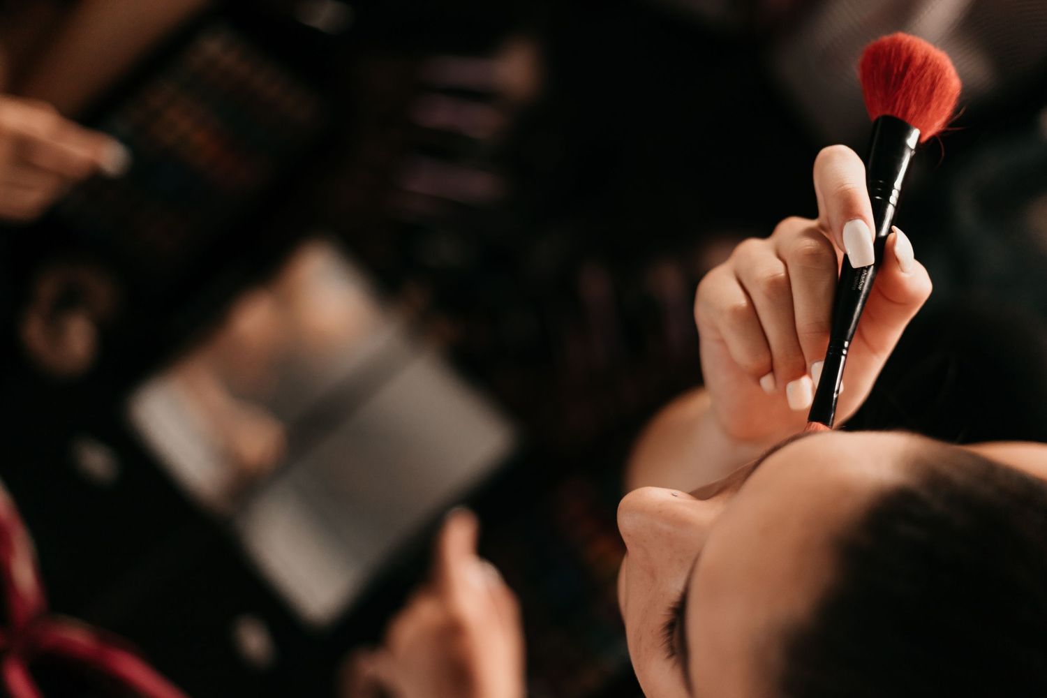 A makeup artist applies blush with a red brush to a model's face in a professional setting.