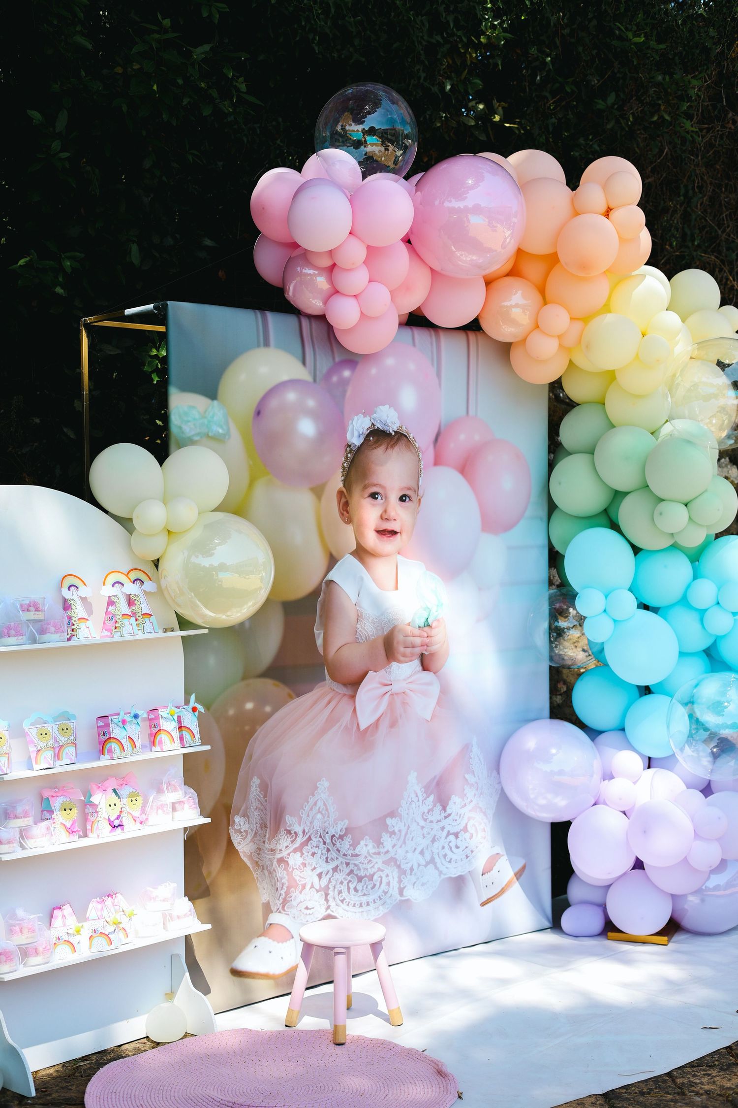 Adorable baby girl in pink dress sitting by colorful rainbow balloon arch at birthday party.