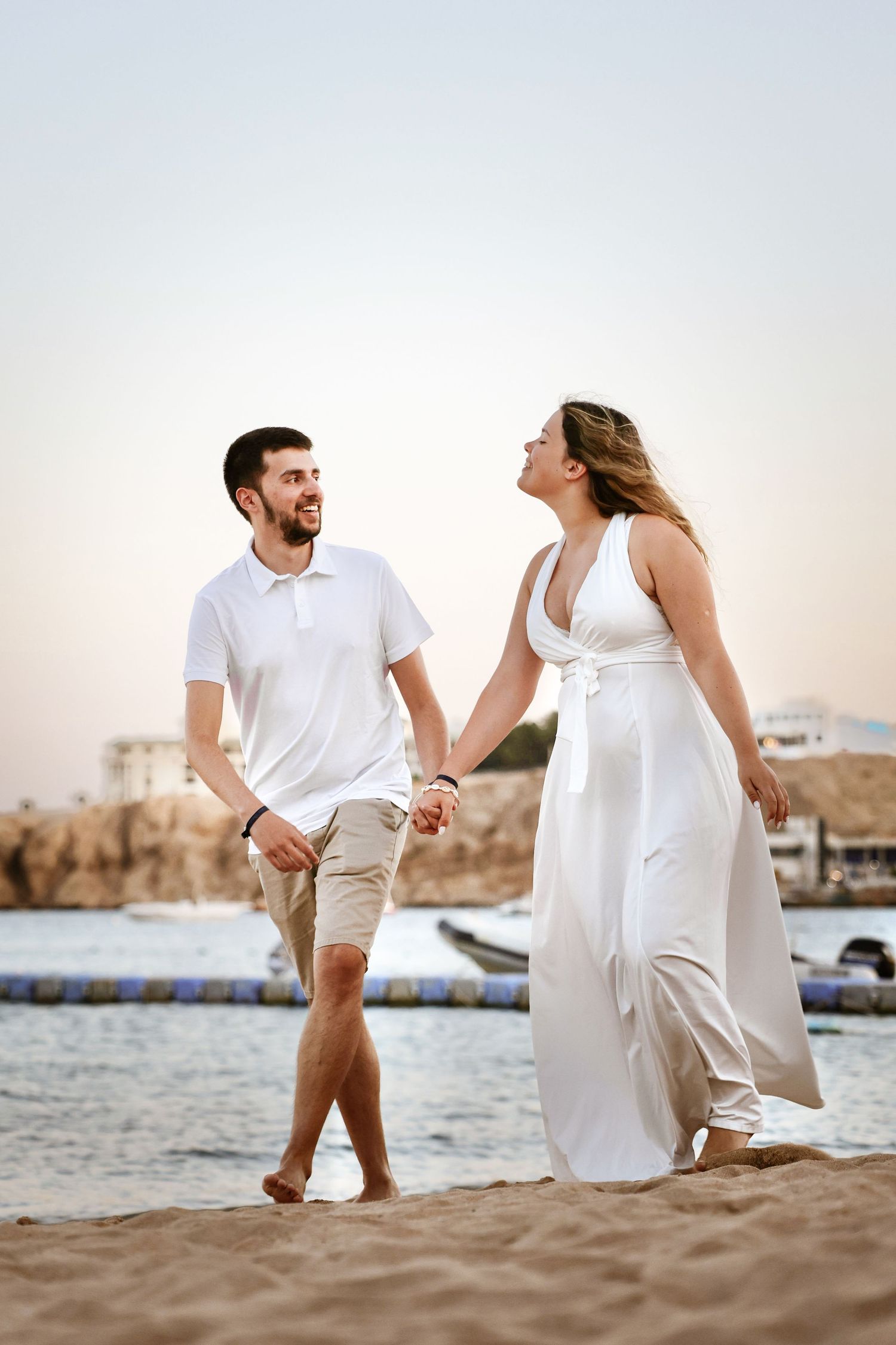 Joyful couple holding hands and strolling barefoot on a sandy beach at sunset.