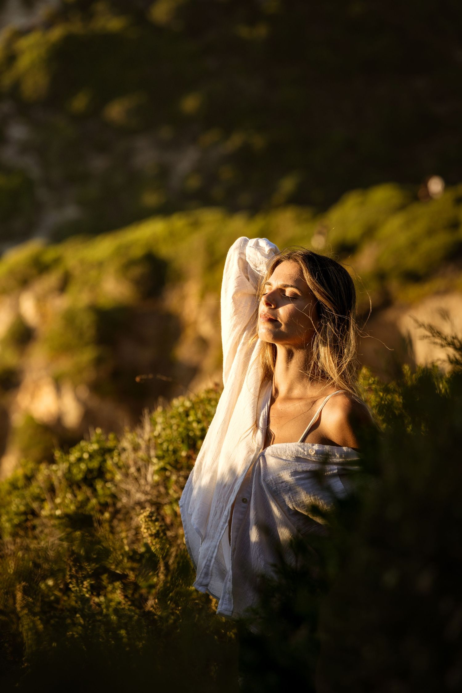 Woman in white dress standing in golden sunset light among green hillside vegetation.