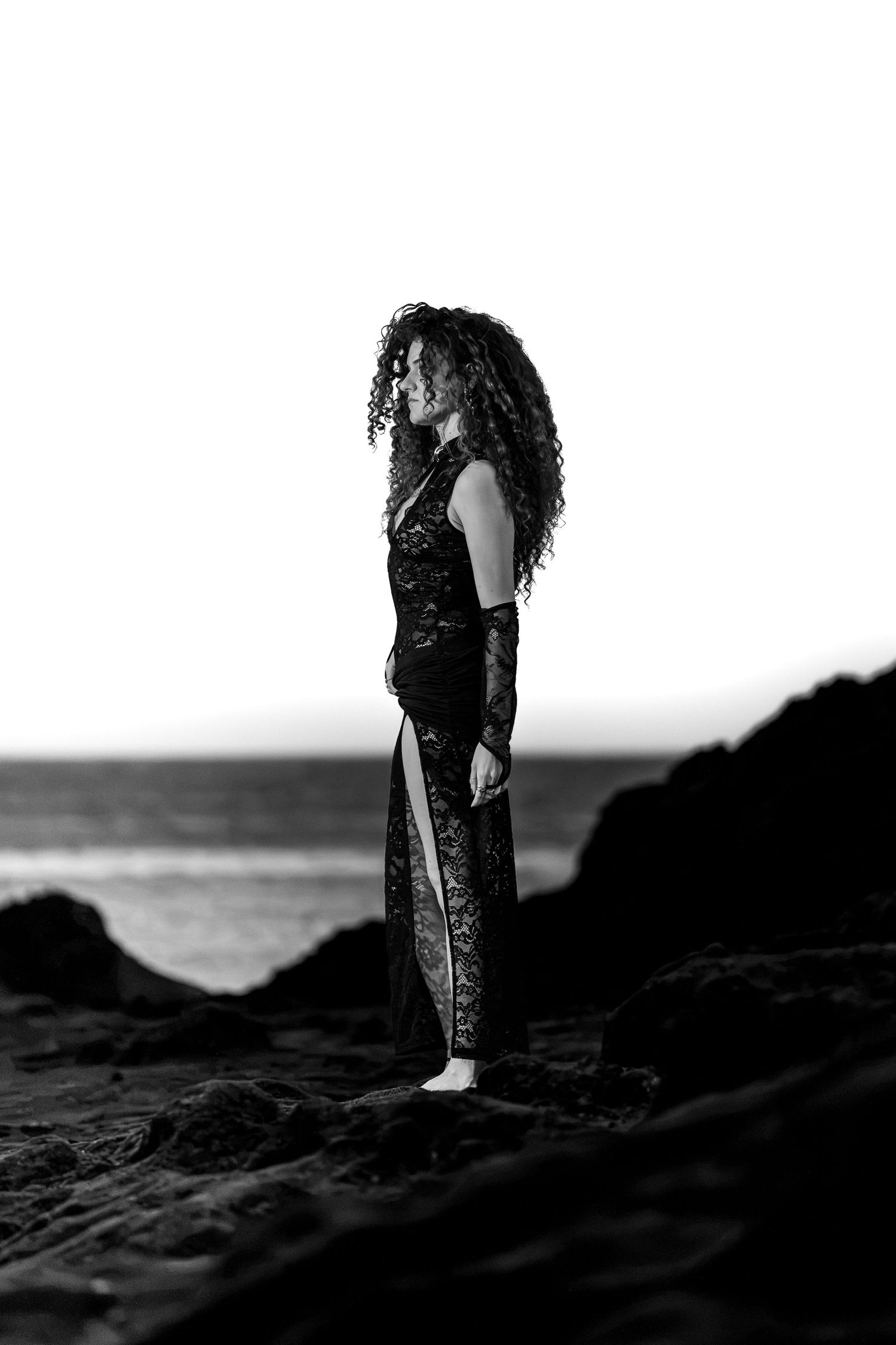 Black and white silhouette of curly-haired woman in black dress standing on beach rocks.