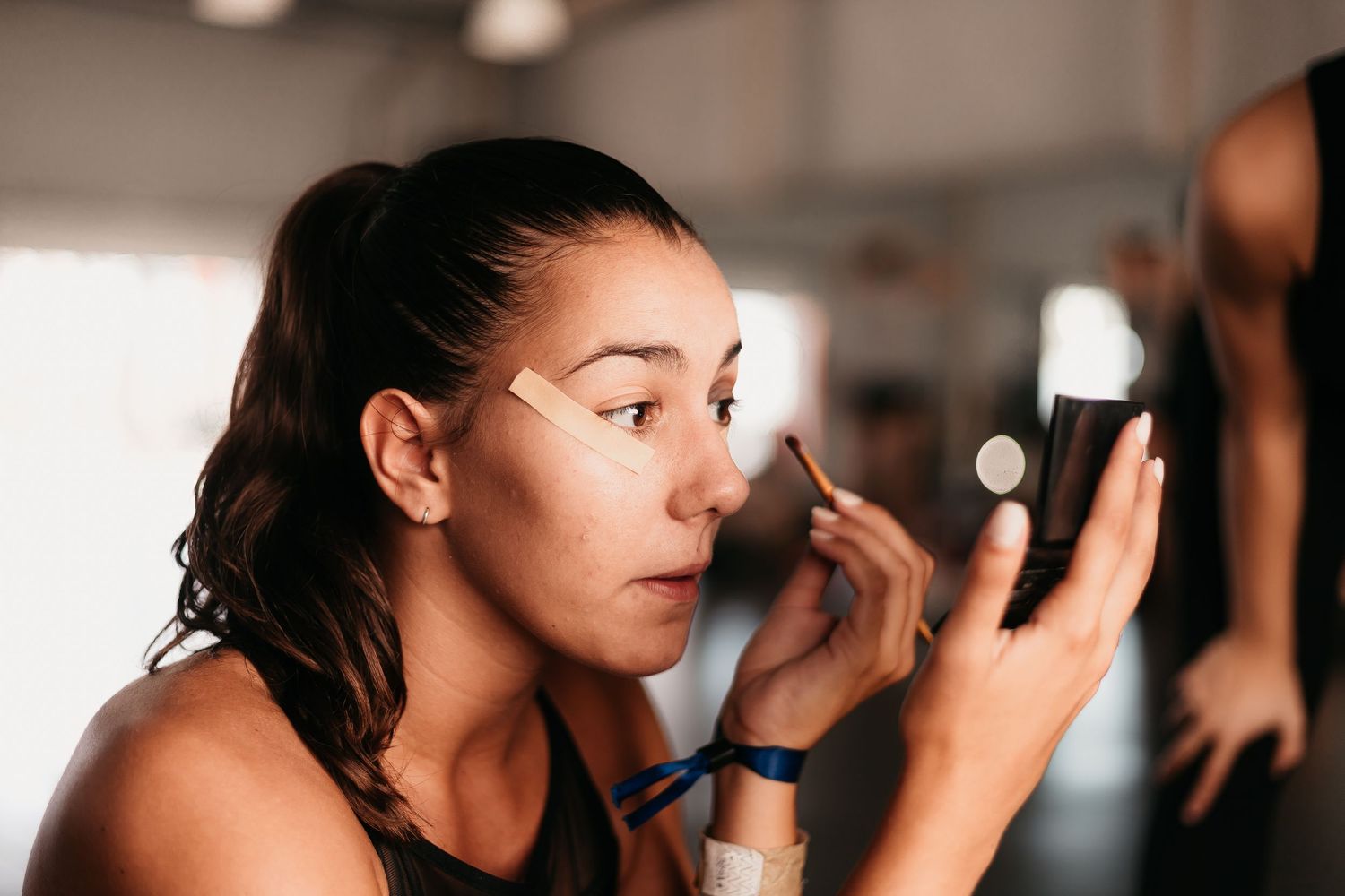 A young woman applies concealer with a brush while looking into a small mirror backstage.