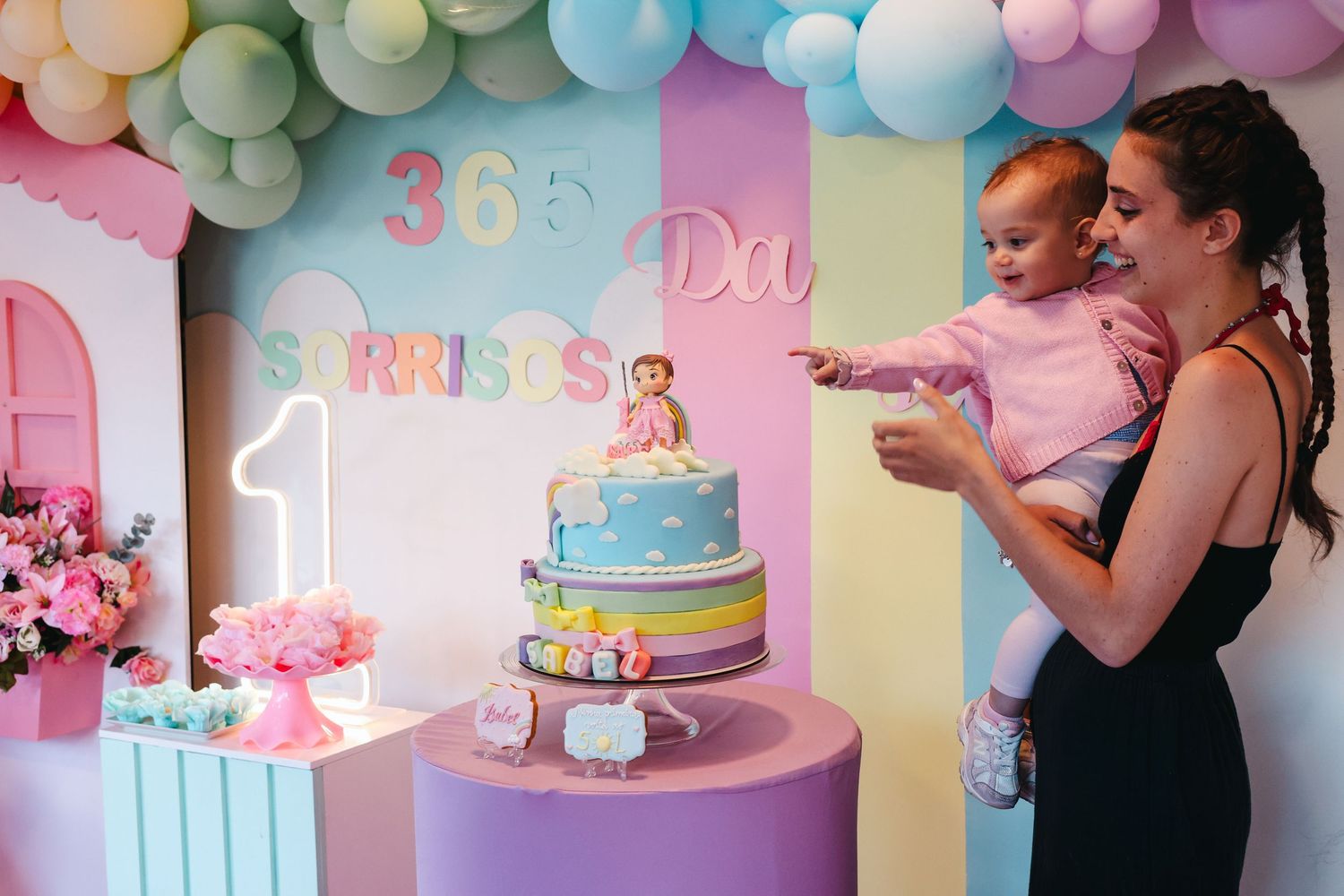 Mother and baby celebrating first birthday with colorful cake and 365 Sorrisos party decoration.