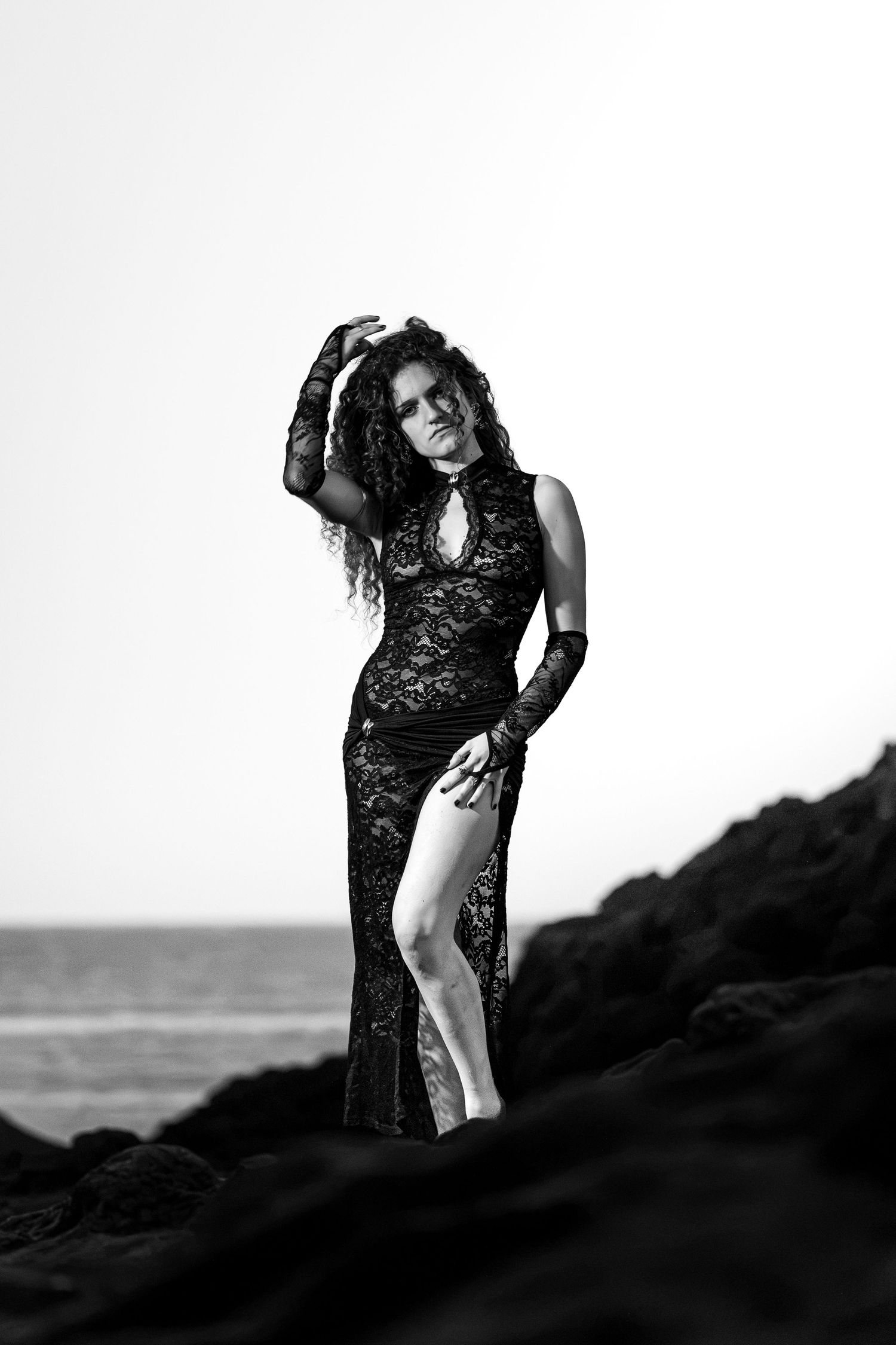 Black and white photo of woman in black lace gown posing on rocks by the ocean.