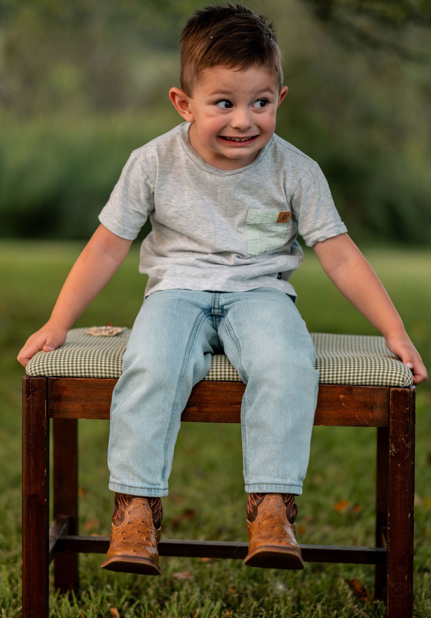 Cheerful toddler boy sitting on a wooden bench outdoors wearing jeans and brown cowboy boots.