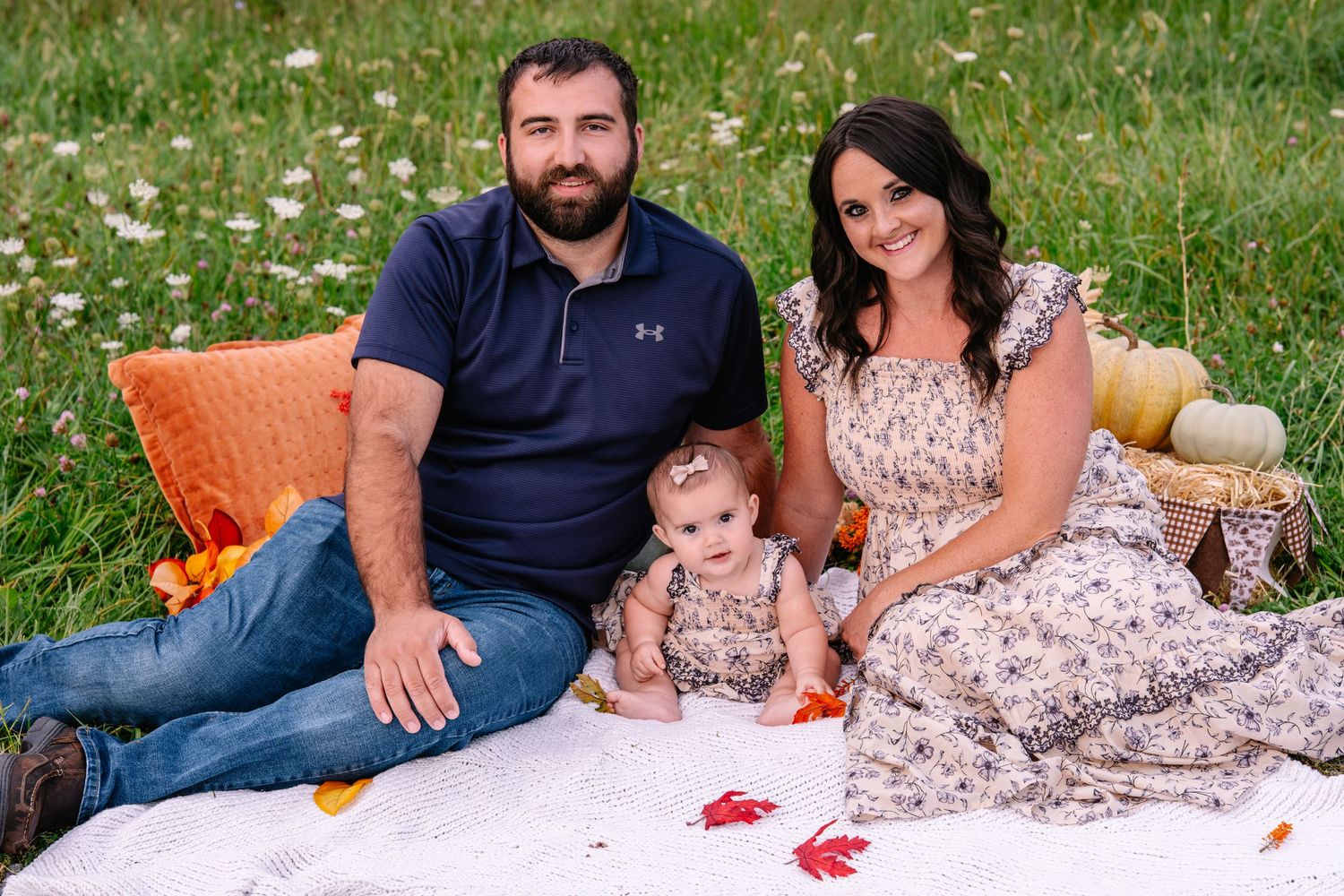 Family of three poses on a white blanket in a flower field, parents smiling with baby girl between them.