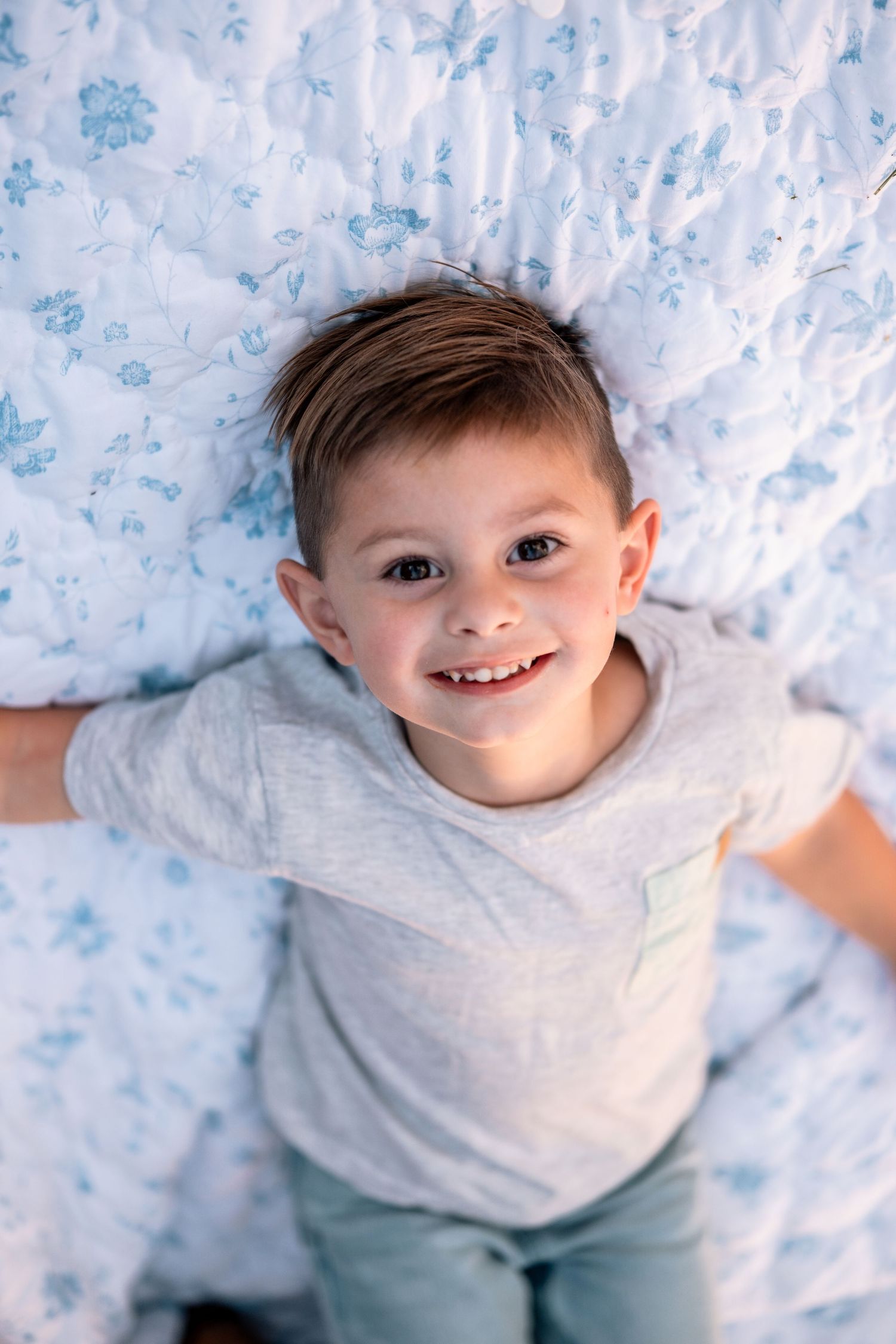 Smiling young boy lying on a floral white and blue bedspread, looking up at the camera cheerfully.
