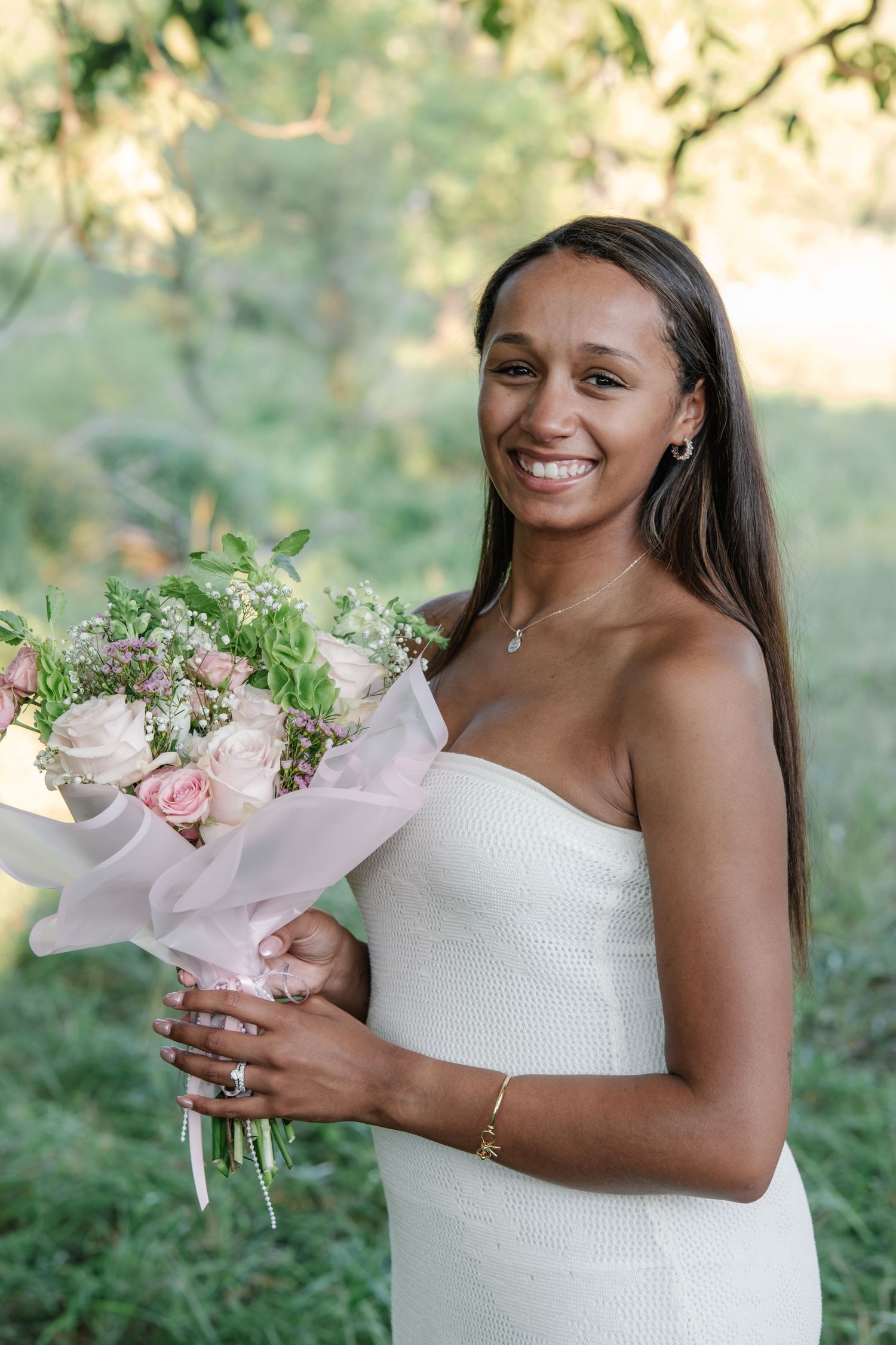 Smiling bride in strapless white dress holding pink rose bouquet outdoors in lush green garden setting.