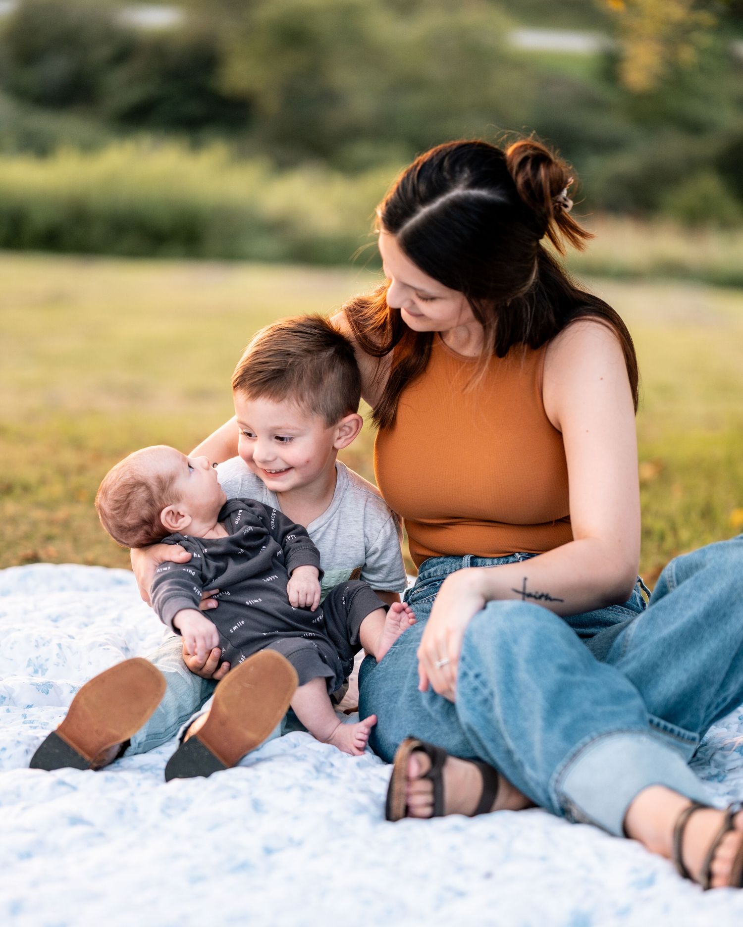 Tender moment as mother in orange top sits with toddler son holding newborn baby on outdoor blanket.
