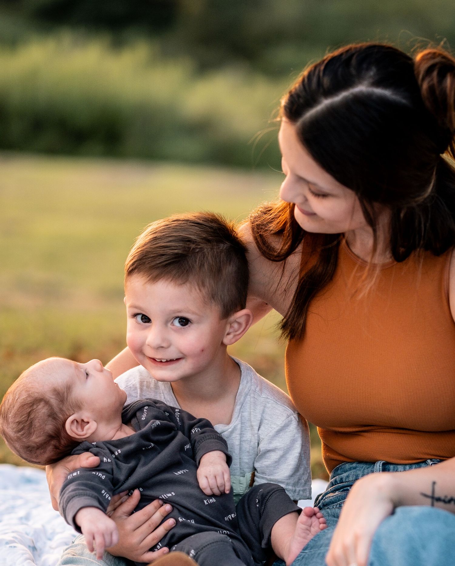 A dark-haired mother in an orange top smiles warmly as her young son holds their newborn sibling outdoors.