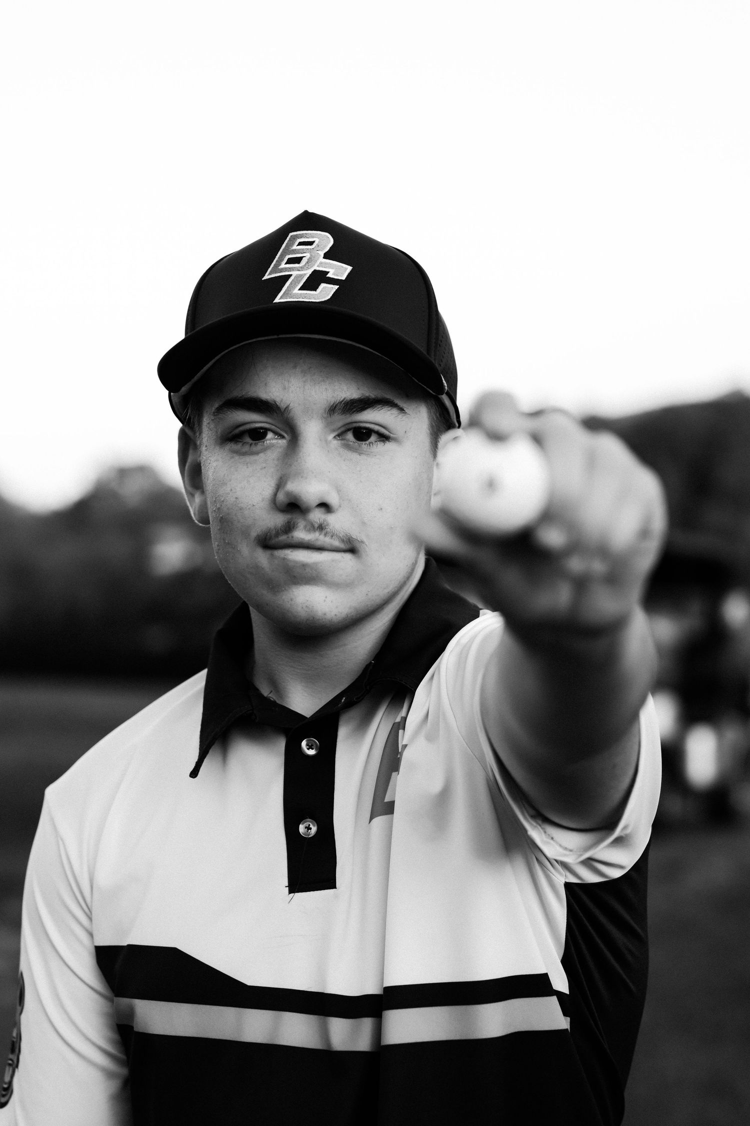 Young golfer in black cap and striped polo holds golf ball toward camera in black and white portrait photo.