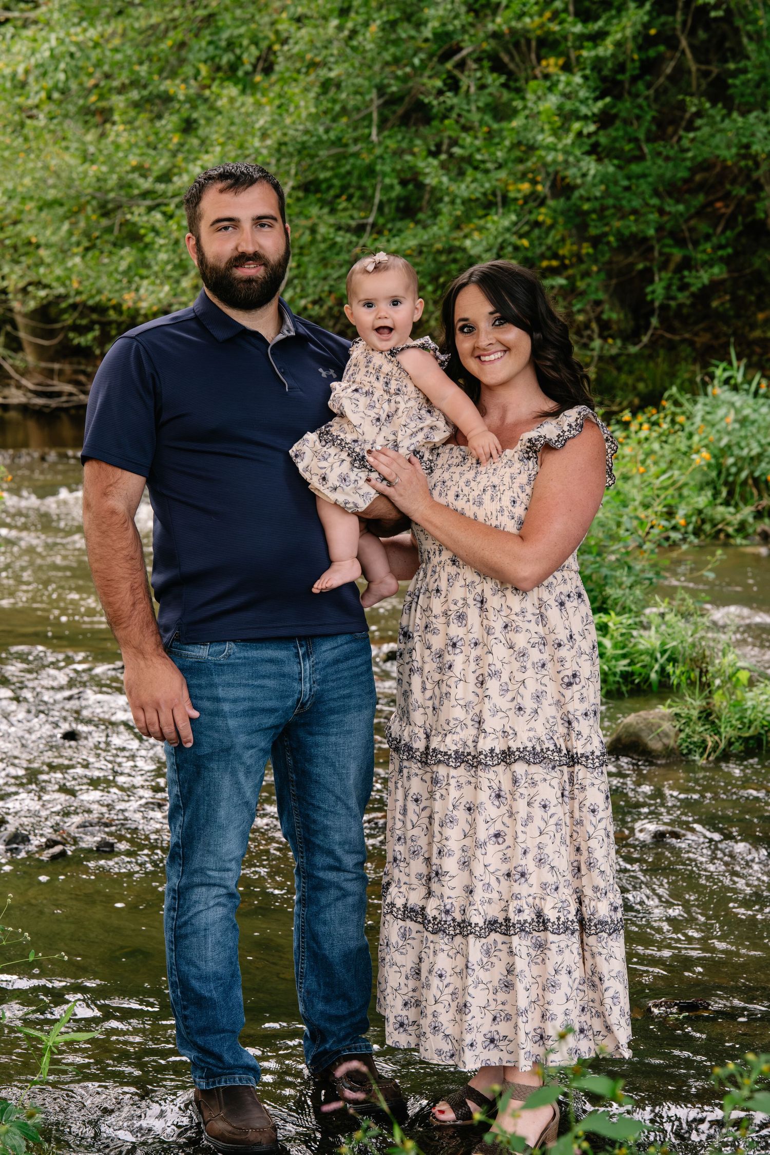 A young family with a bearded man, woman in floral dress, and baby pose by a scenic creek surrounded by greenery.