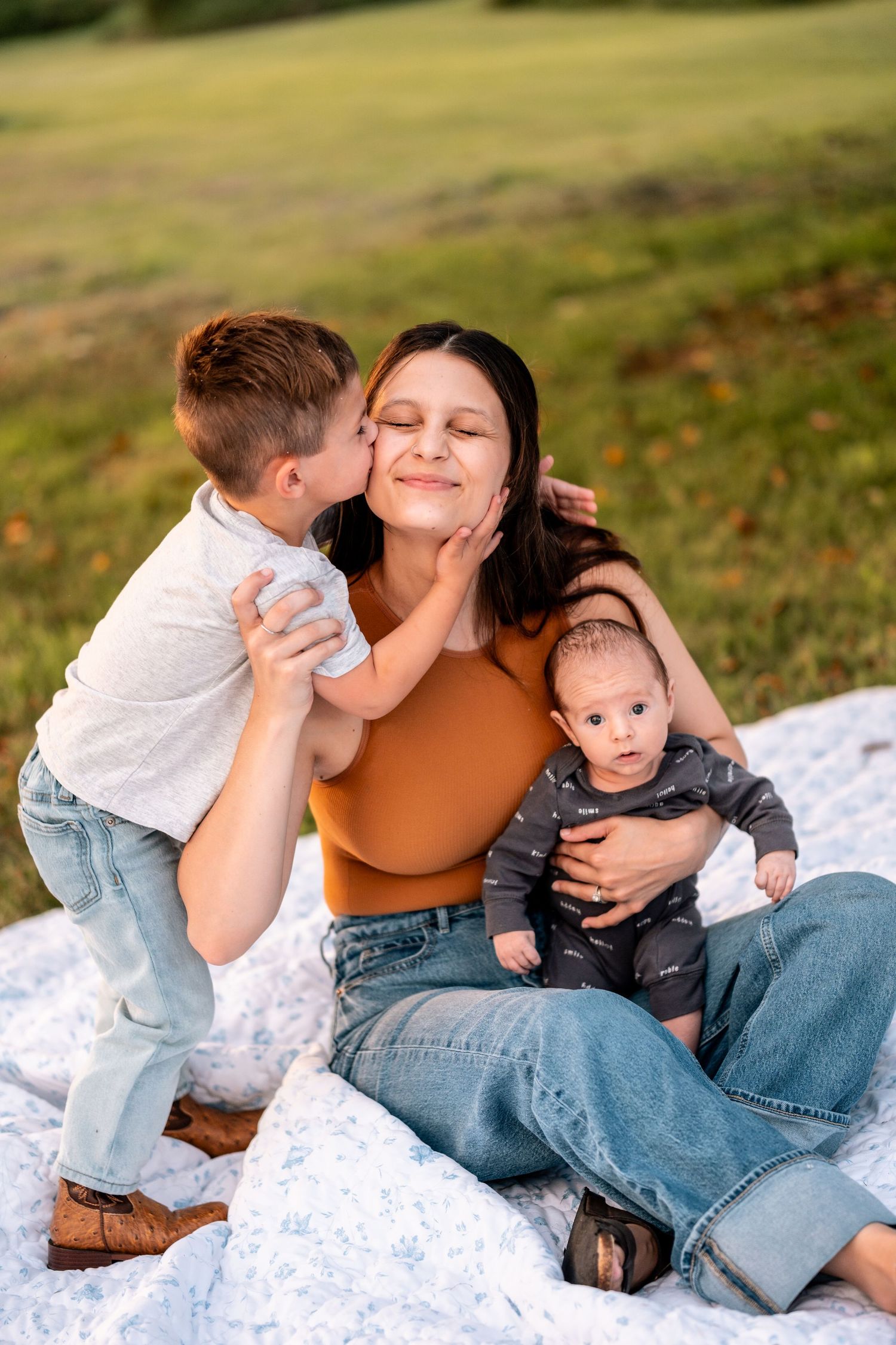 A young boy kisses his smiling mother on the cheek while she holds her baby on a blanket in a green field.