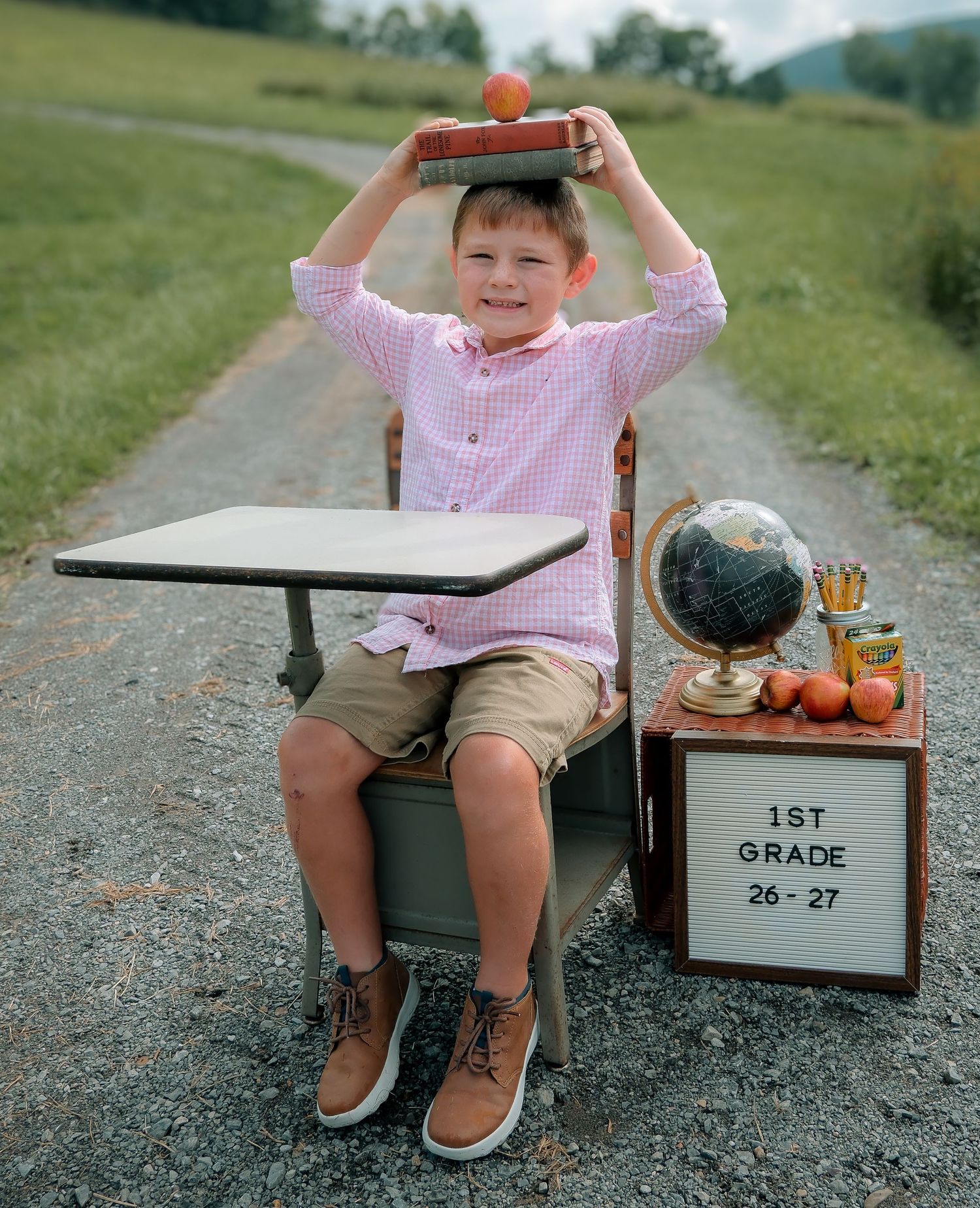 A smiling boy balances books on his head at a vintage school desk outdoors with a 1st grade sign nearby.