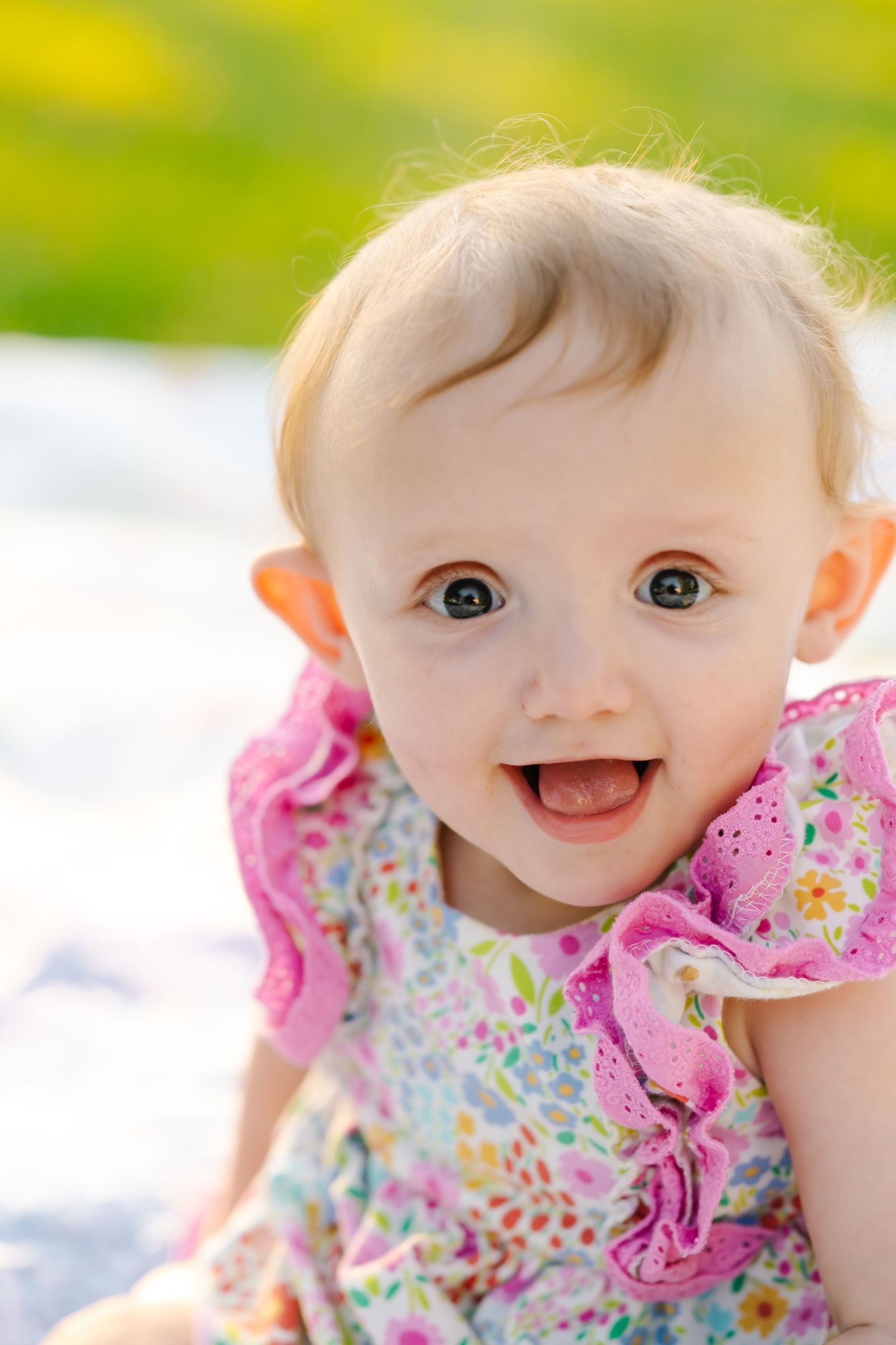 Smiling baby girl in a floral pink dress sitting outdoors on a sunny day with green background.