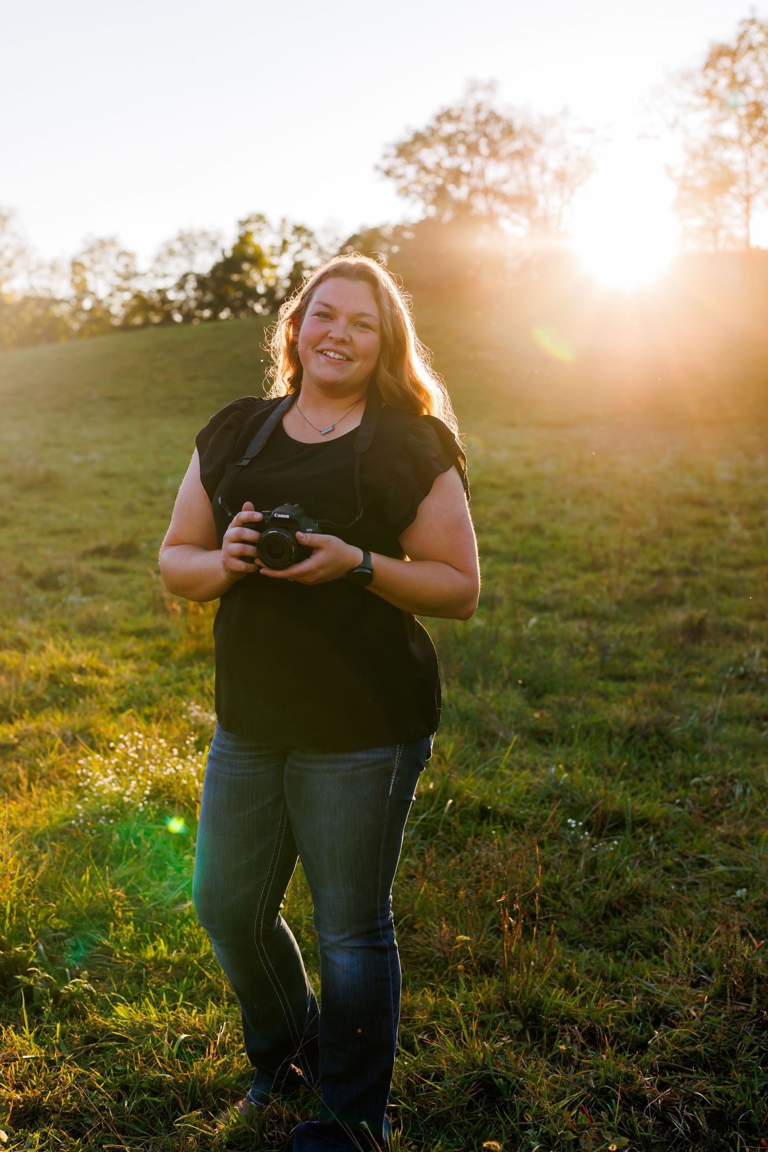 Woman smiling outdoors at golden hour holding a camera in a green field with sunlight glowing behind her.
