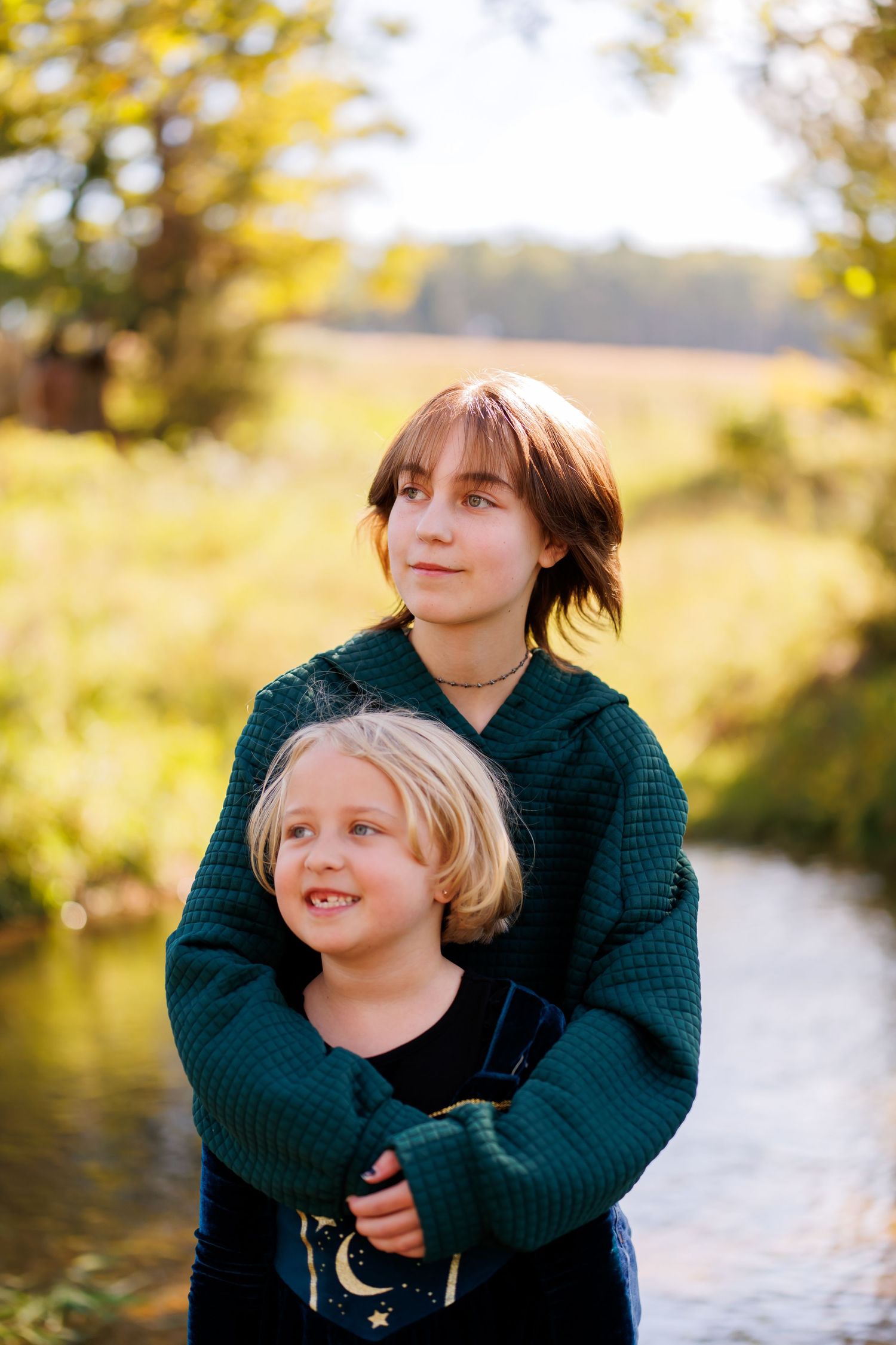 Older child in green sweater hugging younger blonde sibling outdoors near a stream in autumn sunlight.