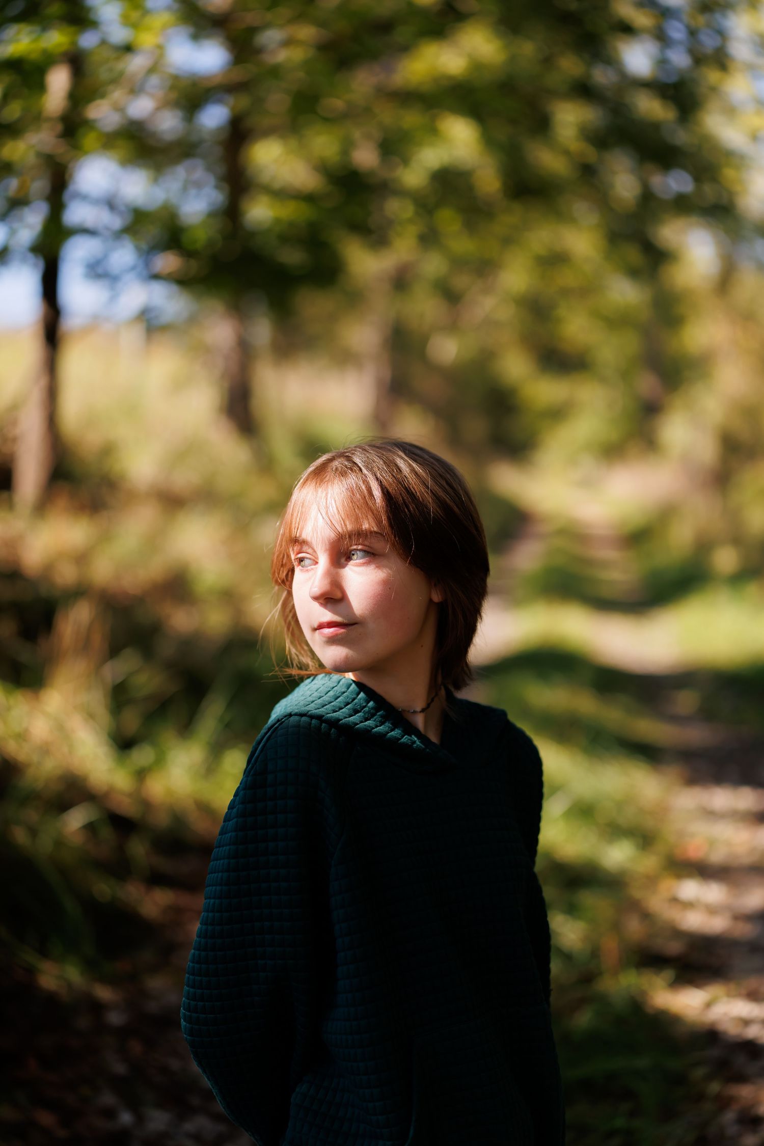 A young woman in a dark green sweater stands on a forest path, gazing upward in soft natural sunlight.