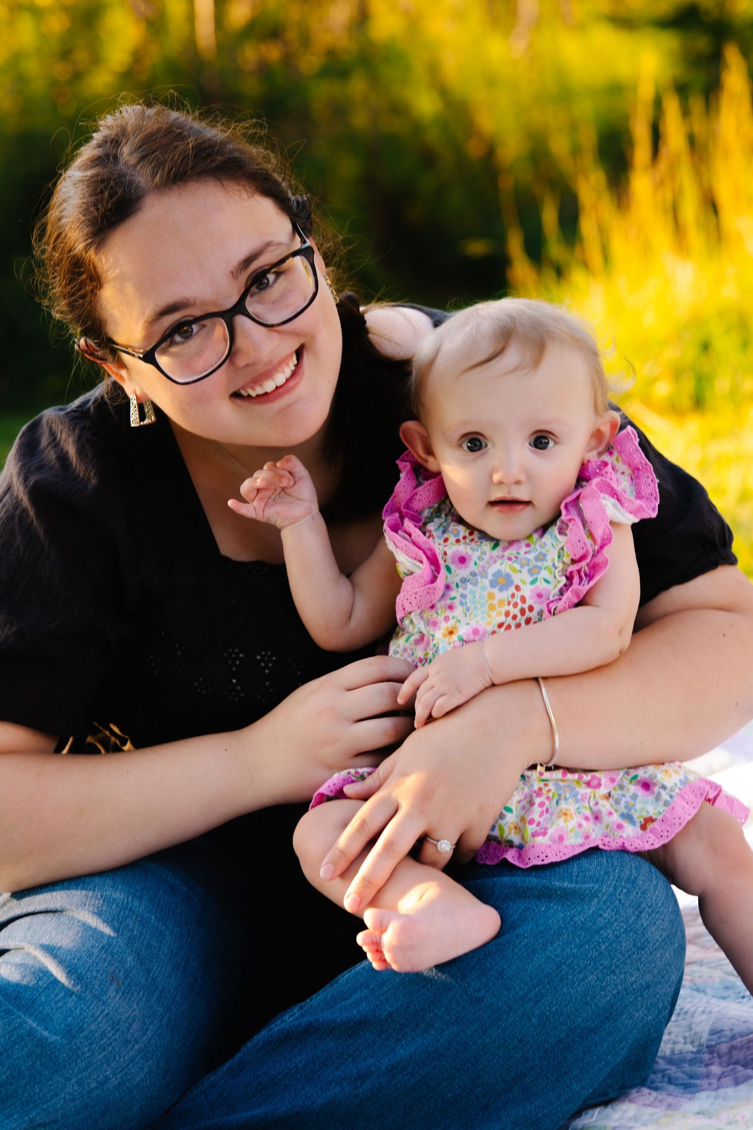 Young woman with glasses smiles holding an adorable baby girl in a floral dress outdoors in golden light.