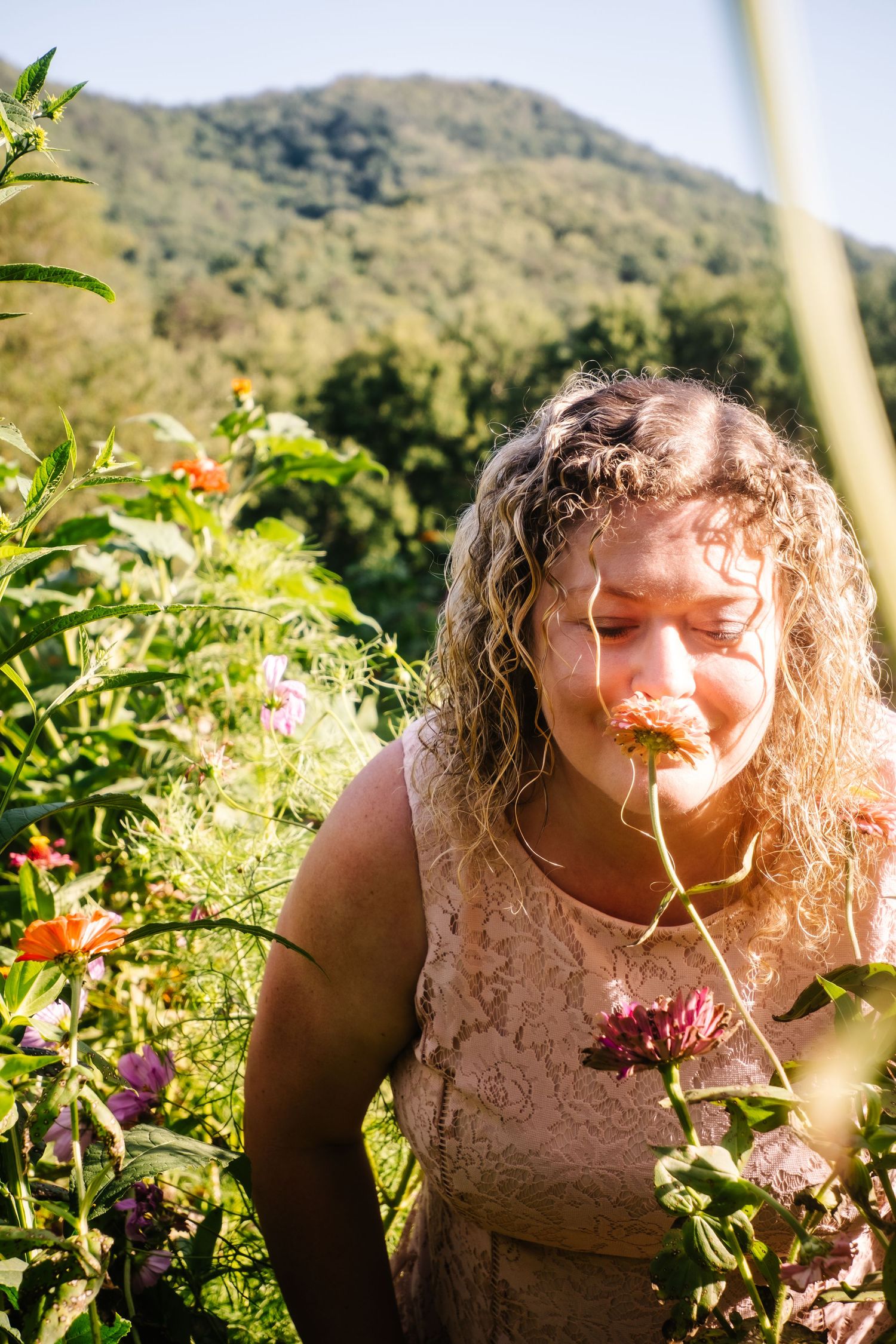 Curly-haired woman smells pink flower in sunny mountain garden landscape.