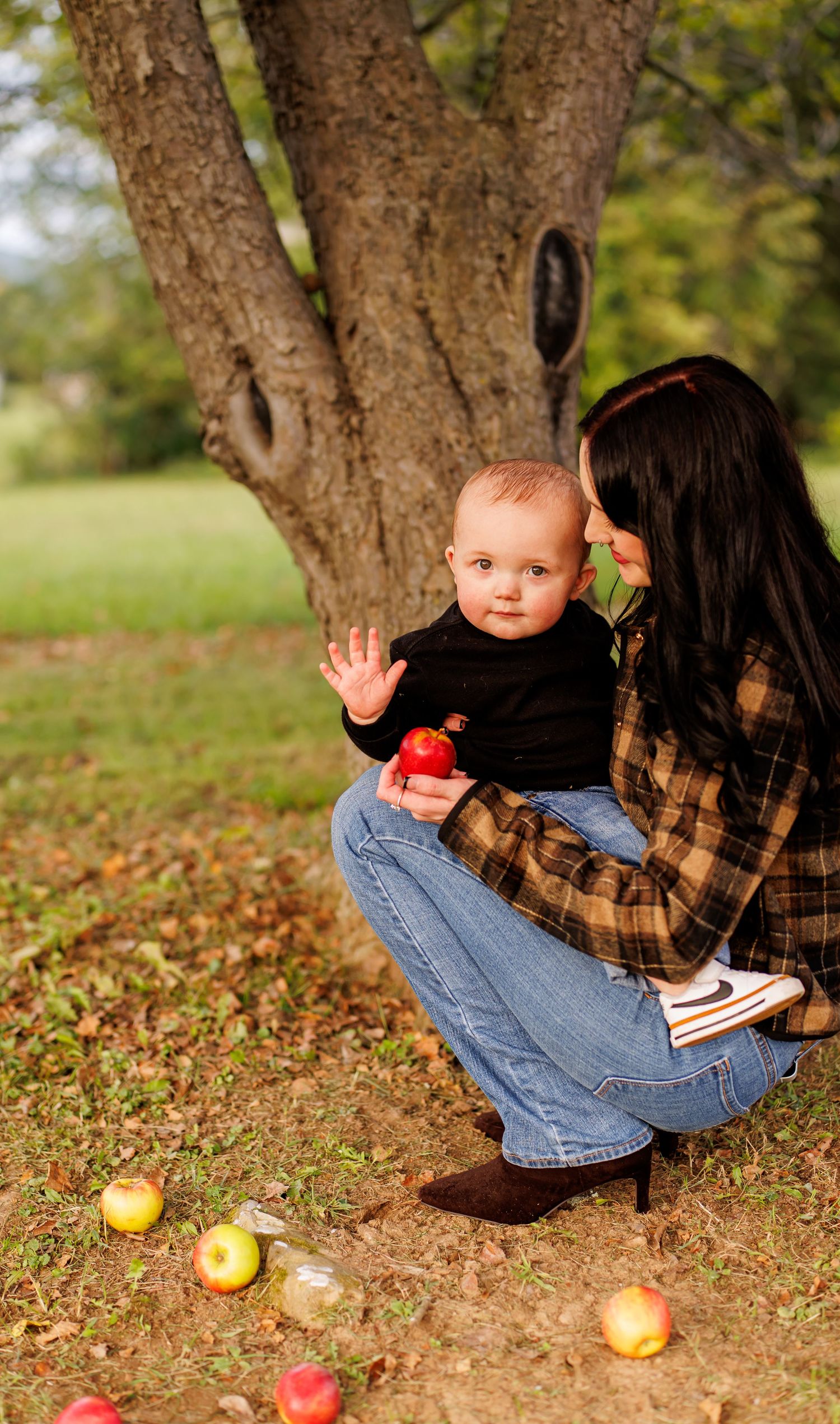 A mother crouches under an apple tree holding her baby who waves while grasping a red apple, with fallen apples on the grass.