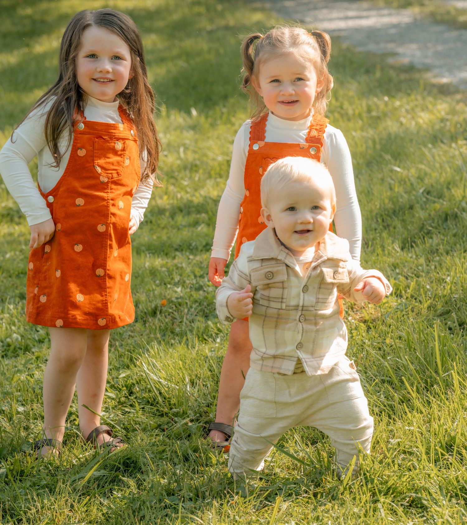 Three young children playing outdoors on green grass, two girls in orange jumpers and a toddler boy in neutral outfit.
