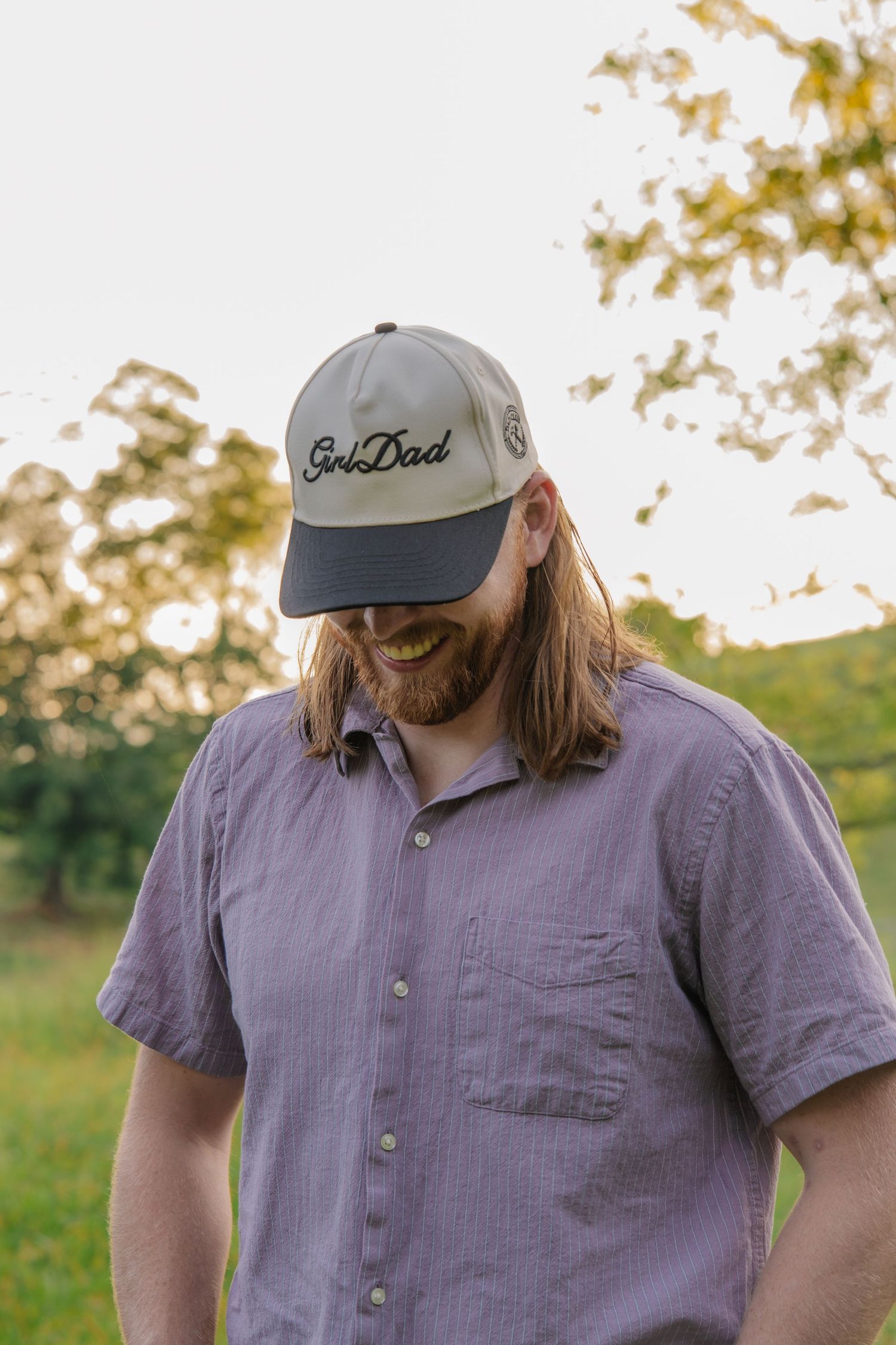 Bearded man with long hair wearing a gray Golf Dad cap and purple shirt outdoors at golden hour.