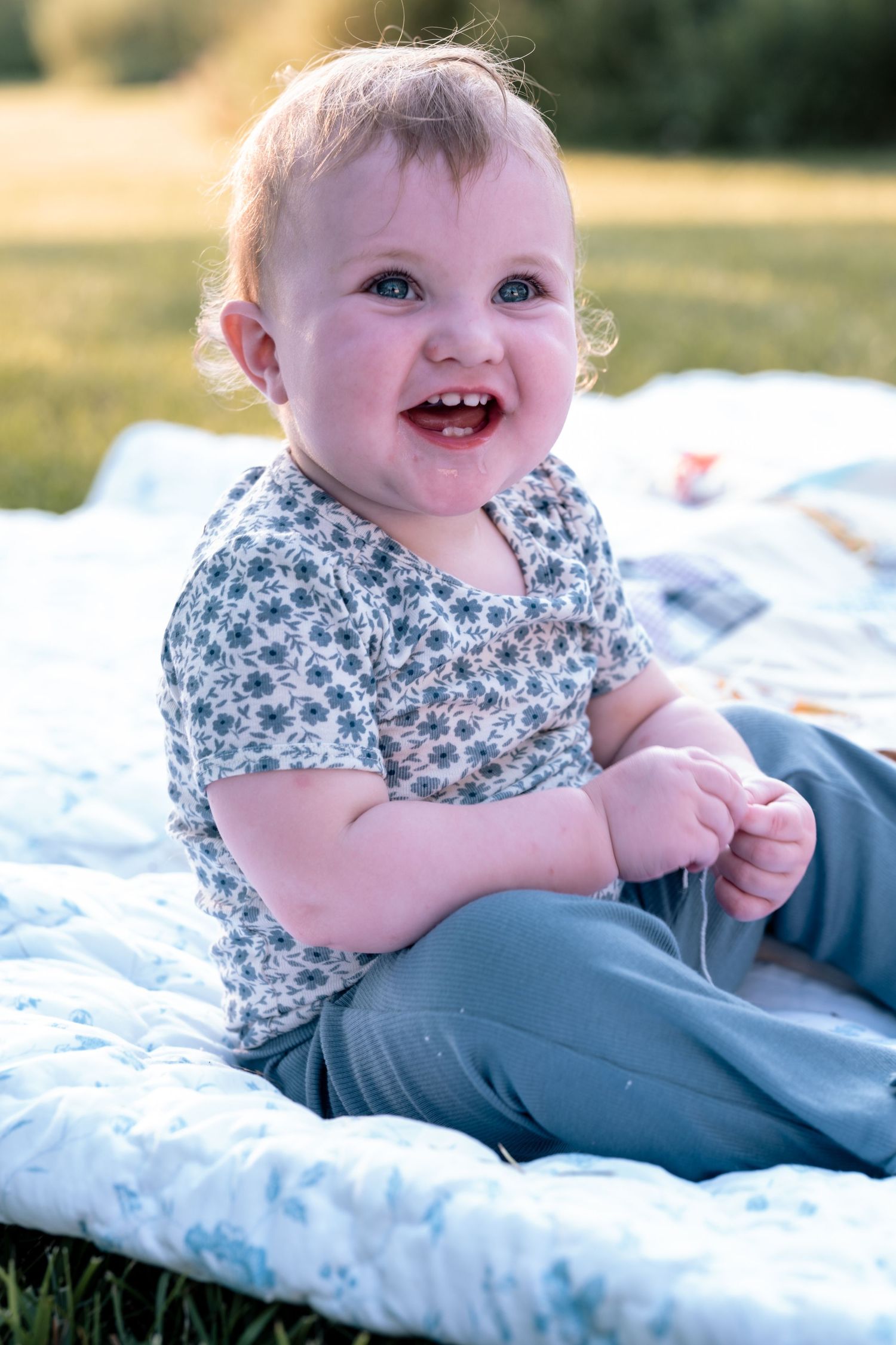 Happy baby sitting on a floral quilt outdoors, smiling brightly in golden sunlight wearing a floral shirt and gray pants.