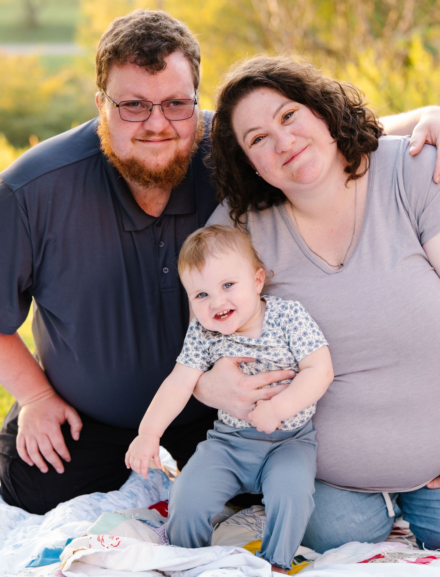 A happy family of three poses outdoors, with parents smiling alongside their cheerful toddler on a blanket.