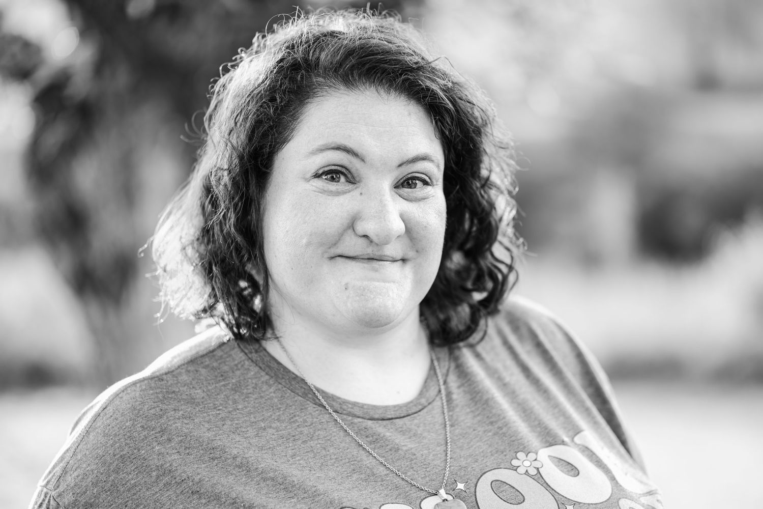 Black and white portrait of a smiling woman with curly hair wearing a casual t-shirt outdoors.