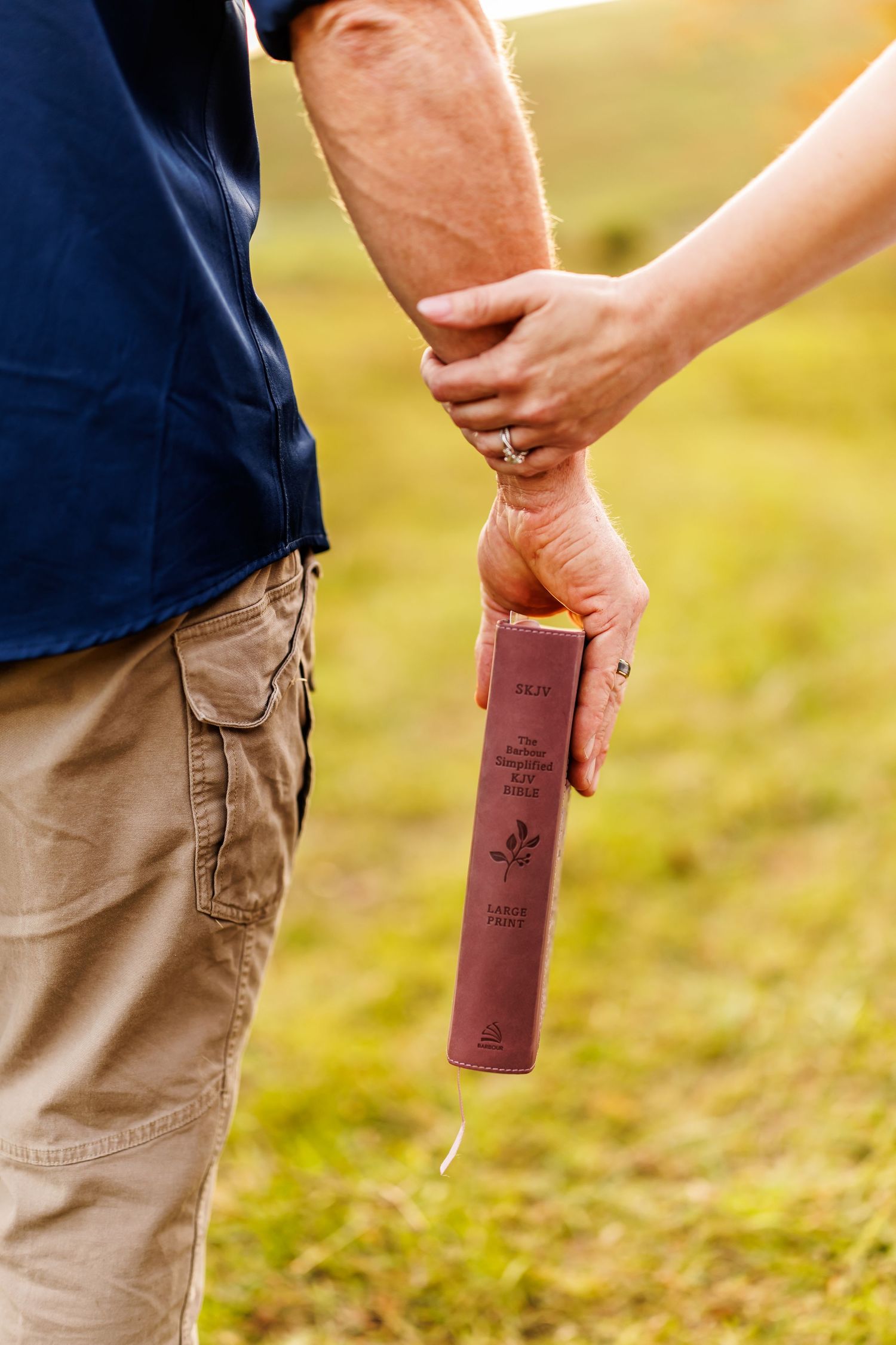 Couple holding hands outdoors while man carries a brown leather Holy Bible in a sunlit green field.